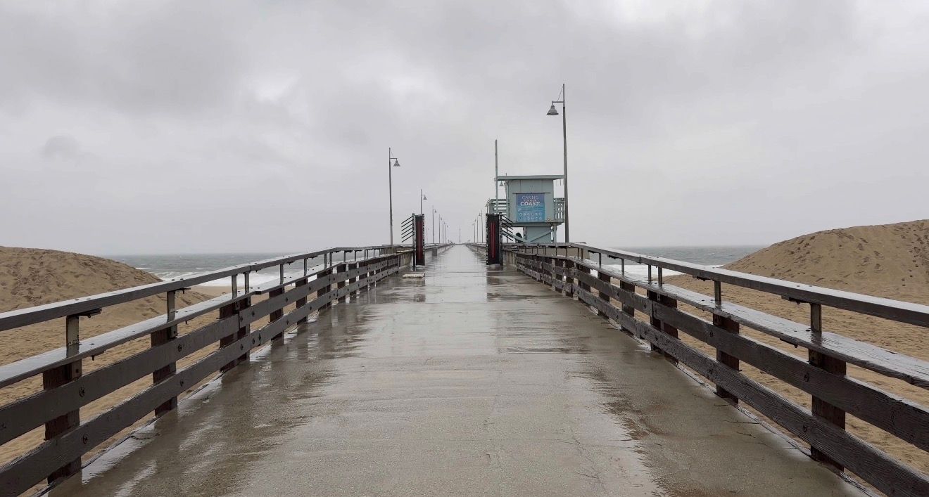 A foggy wooden pier extends into the ocean with waves crashing against it during an overcast day.
