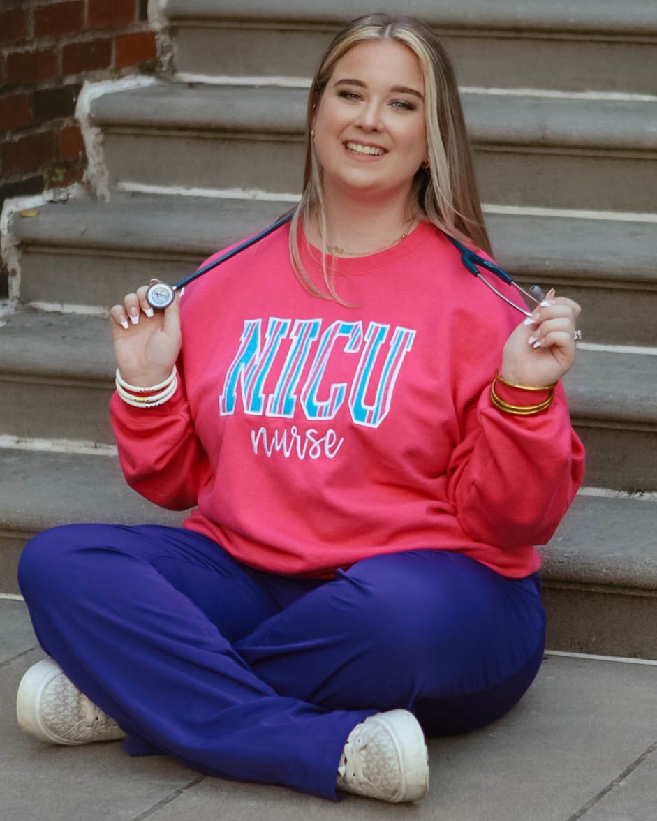 Student wearing pink NICU nurse sweatshirt and blue pants sitting on concrete steps.