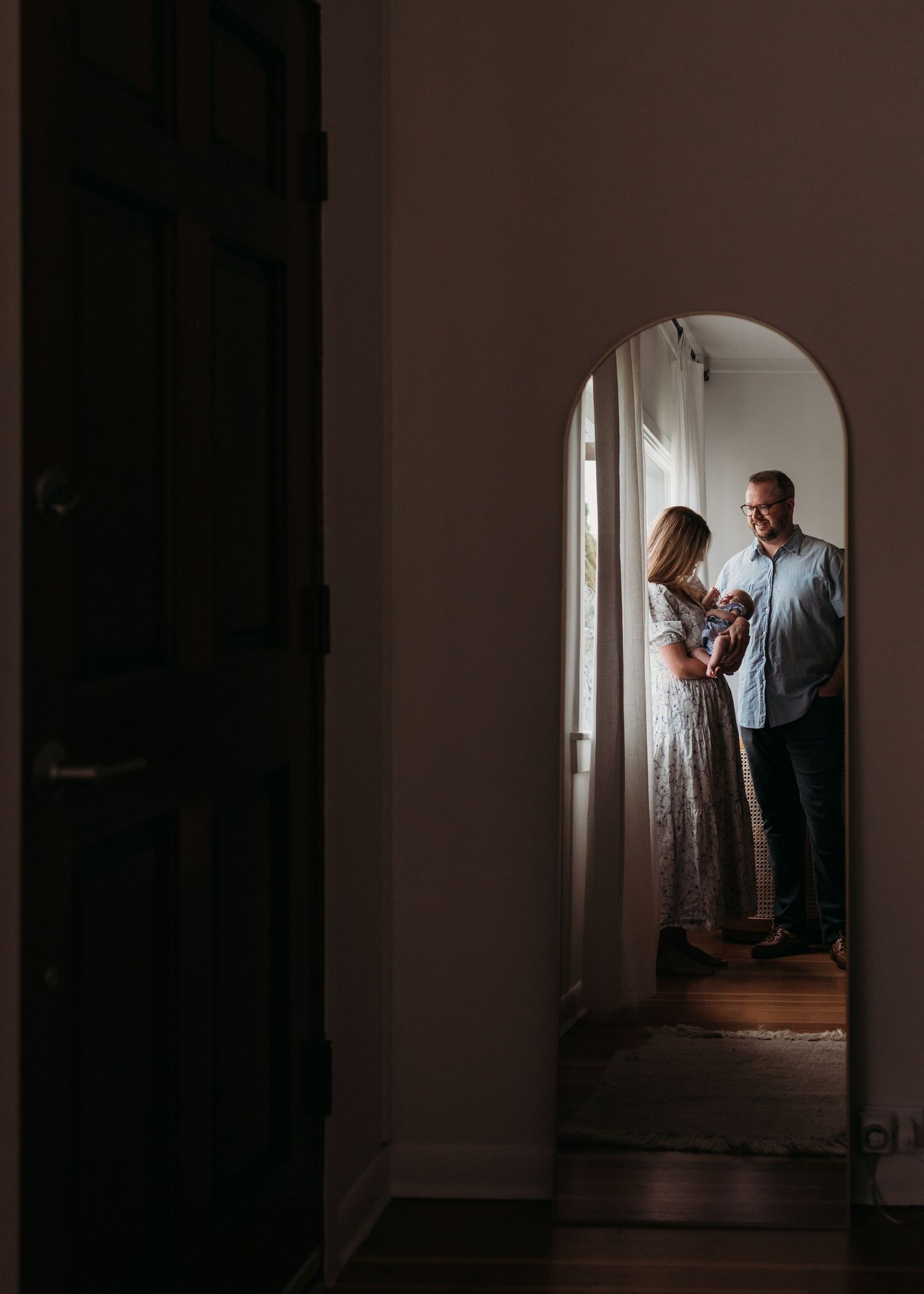A couple stands in conversation through an arched doorway, silhouetted by natural window light in a moody interior.