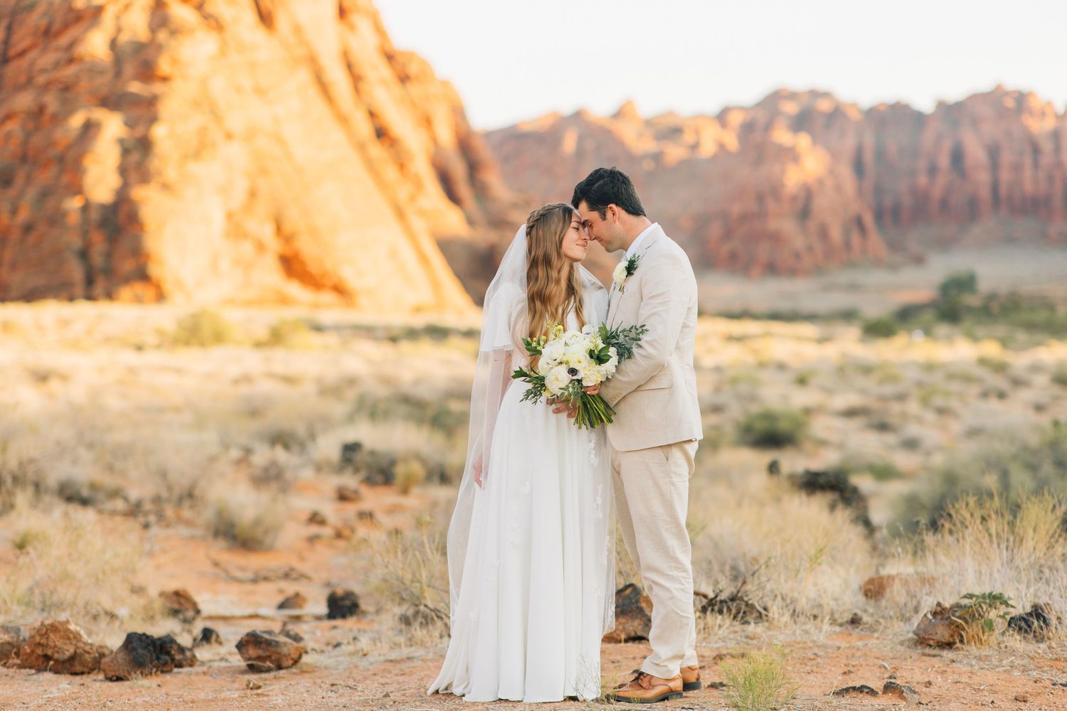 Newlywed couple shares romantic moments against stunning red rock formations in a desert landscape at sunset.
