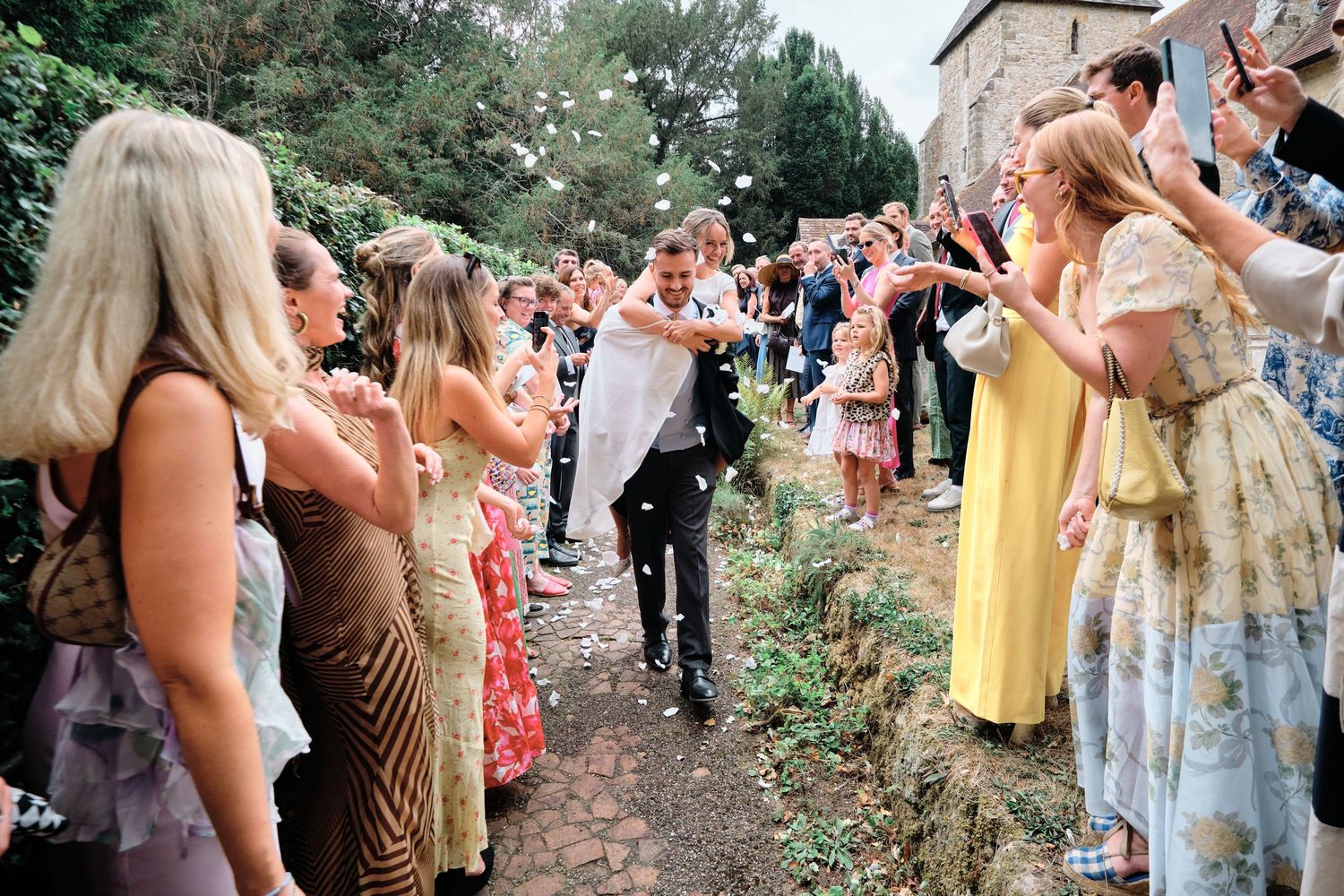 Wedding guests line a church path while celebrating and throwing confetti at the happy couple during an outdoor celebration.