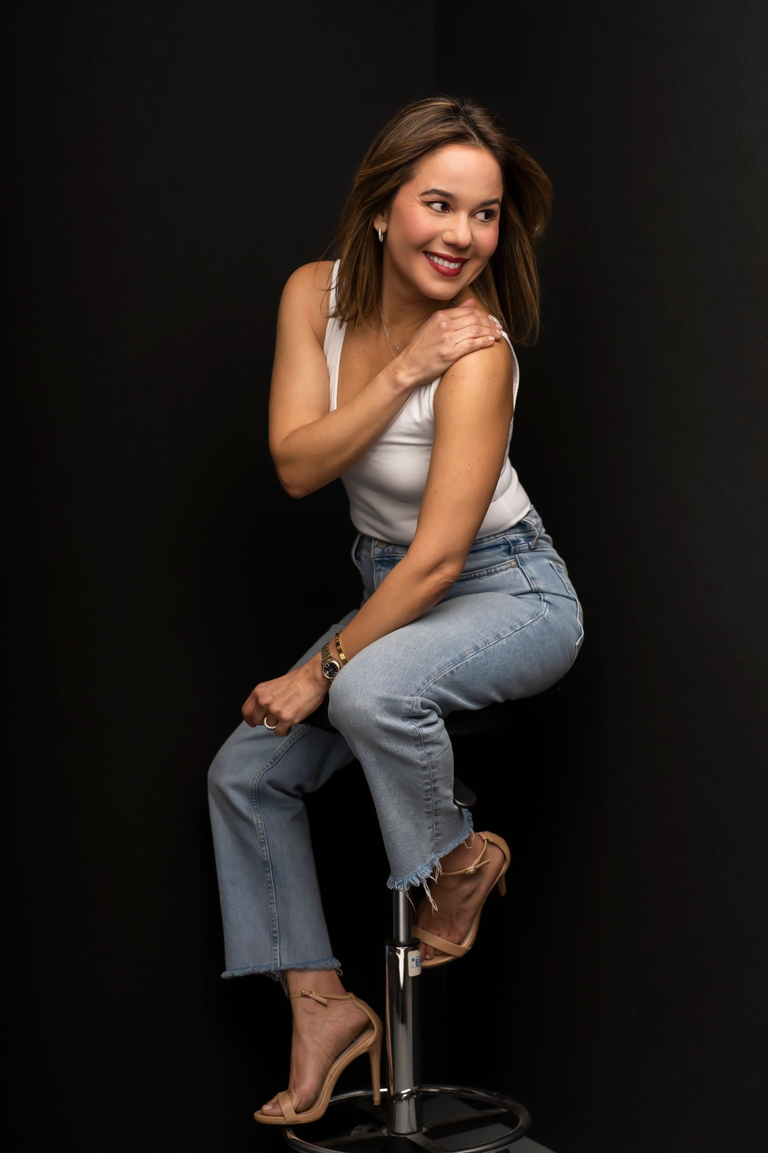 Studio portrait of person in white top and jeans posed on stool against black background.