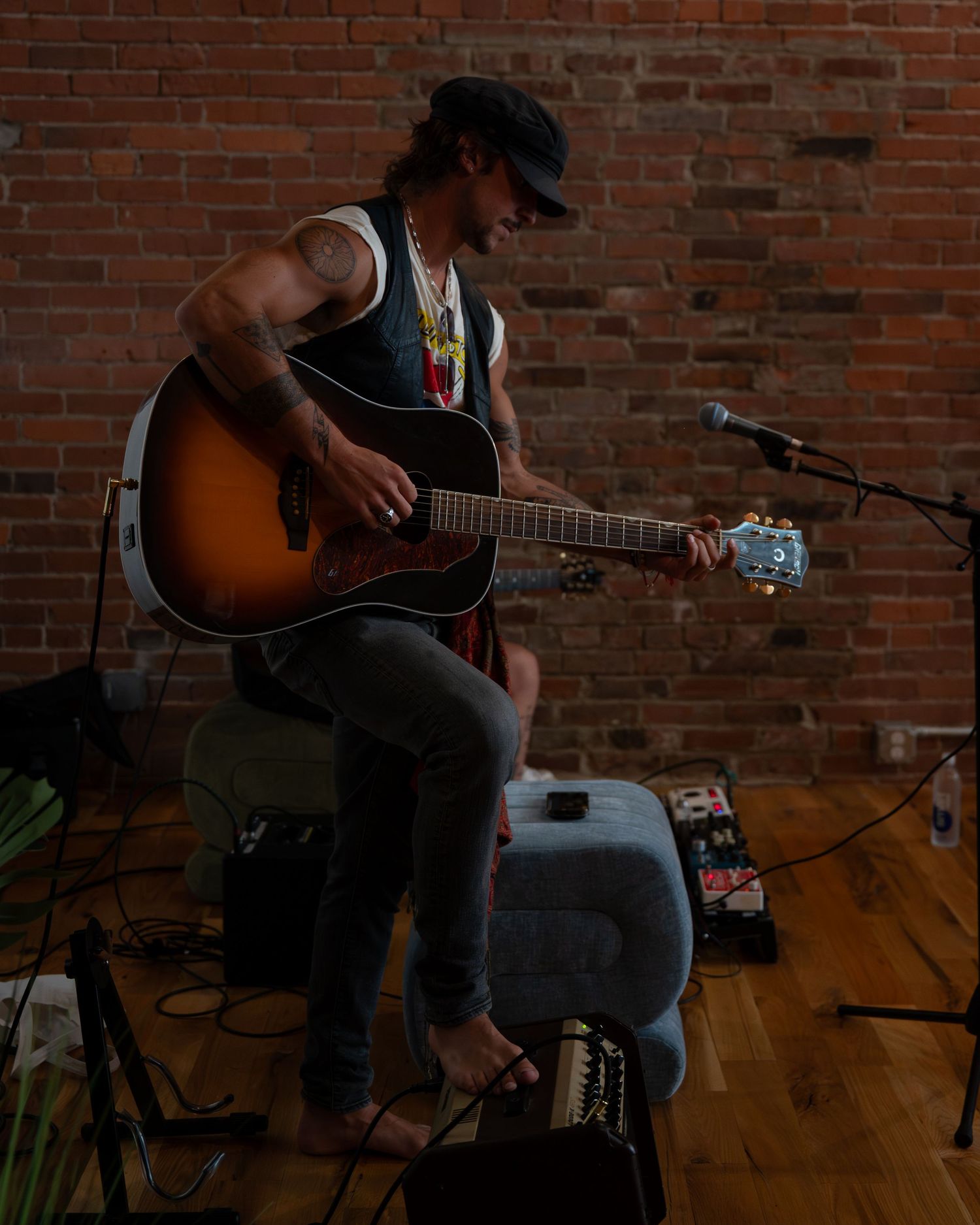 A musician performs with an acoustic guitar against a rustic brick wall backdrop in moody lighting.