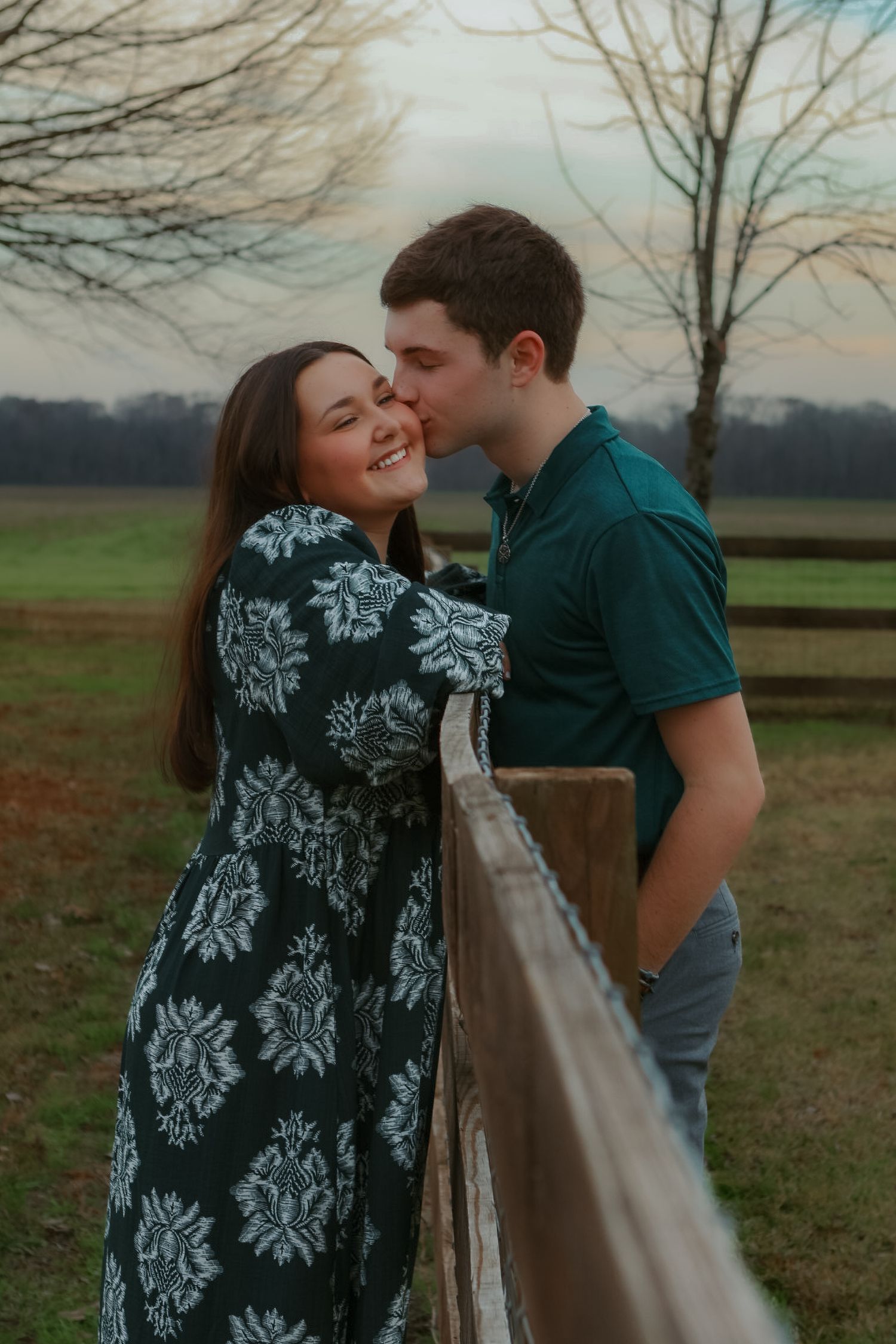 A couple shares romantic moments by a rustic wooden fence in a pastoral countryside setting at sunset.