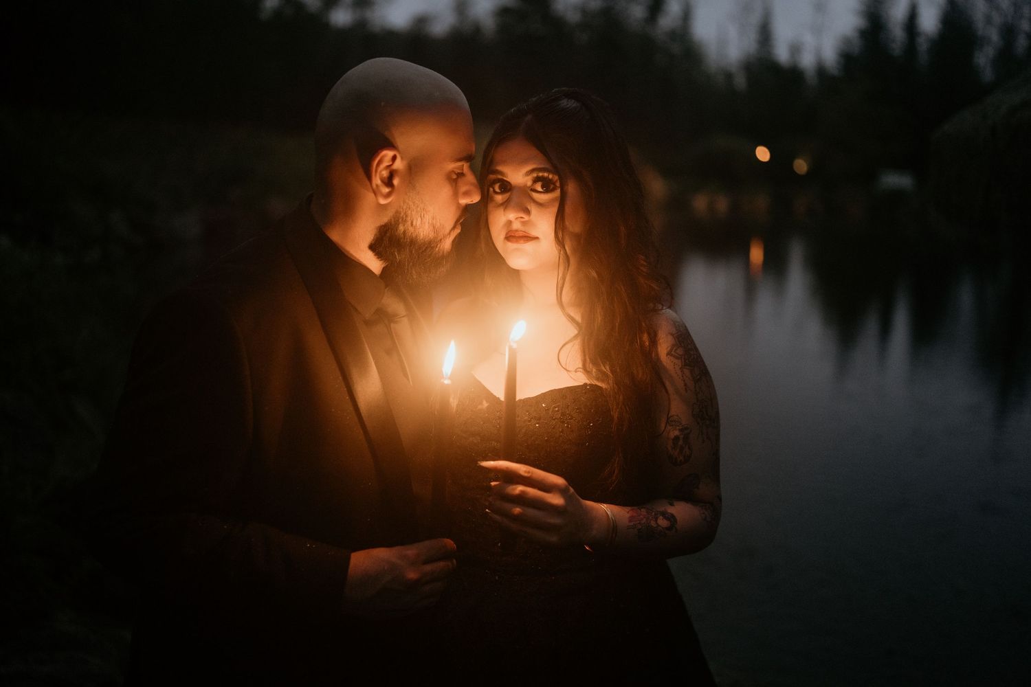 A romantic couple illuminated by candlelight stands beside a dark lake at dusk, creating an intimate atmosphere.