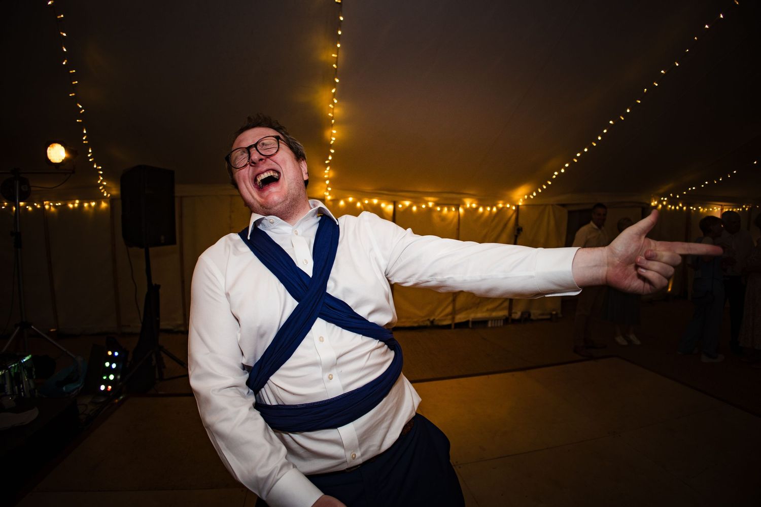 A dad with a baby carrier dances joyfully under string lights at a party tent in Nova Scotia wedding.