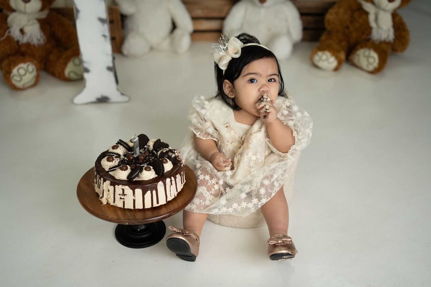 Toddler in frilly cream outfit sits next to chocolate drip cake while surrounded by plush stuffed animals.
