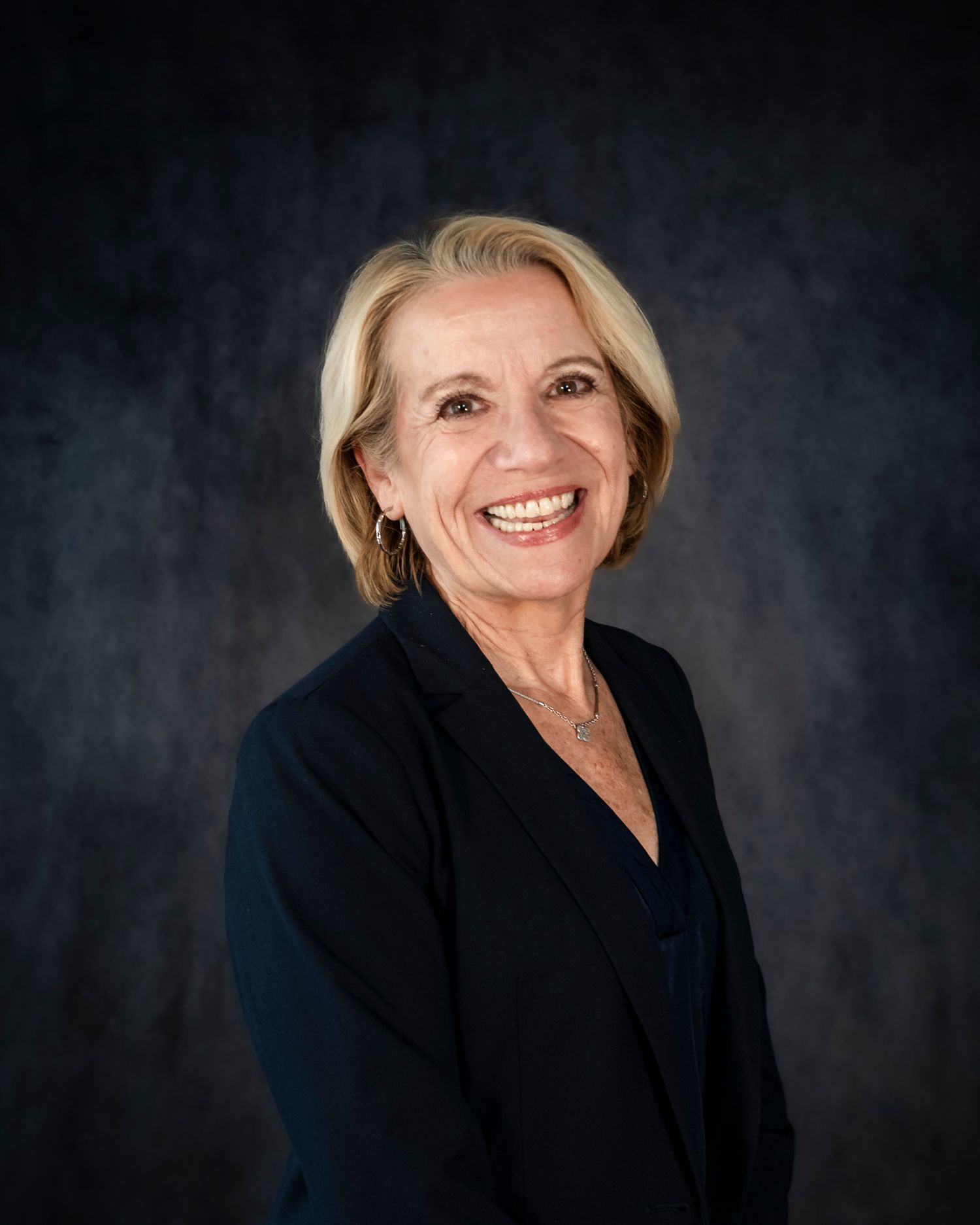 Professional headshot portrait of someone in a black blazer smiling warmly against a dark gray background.