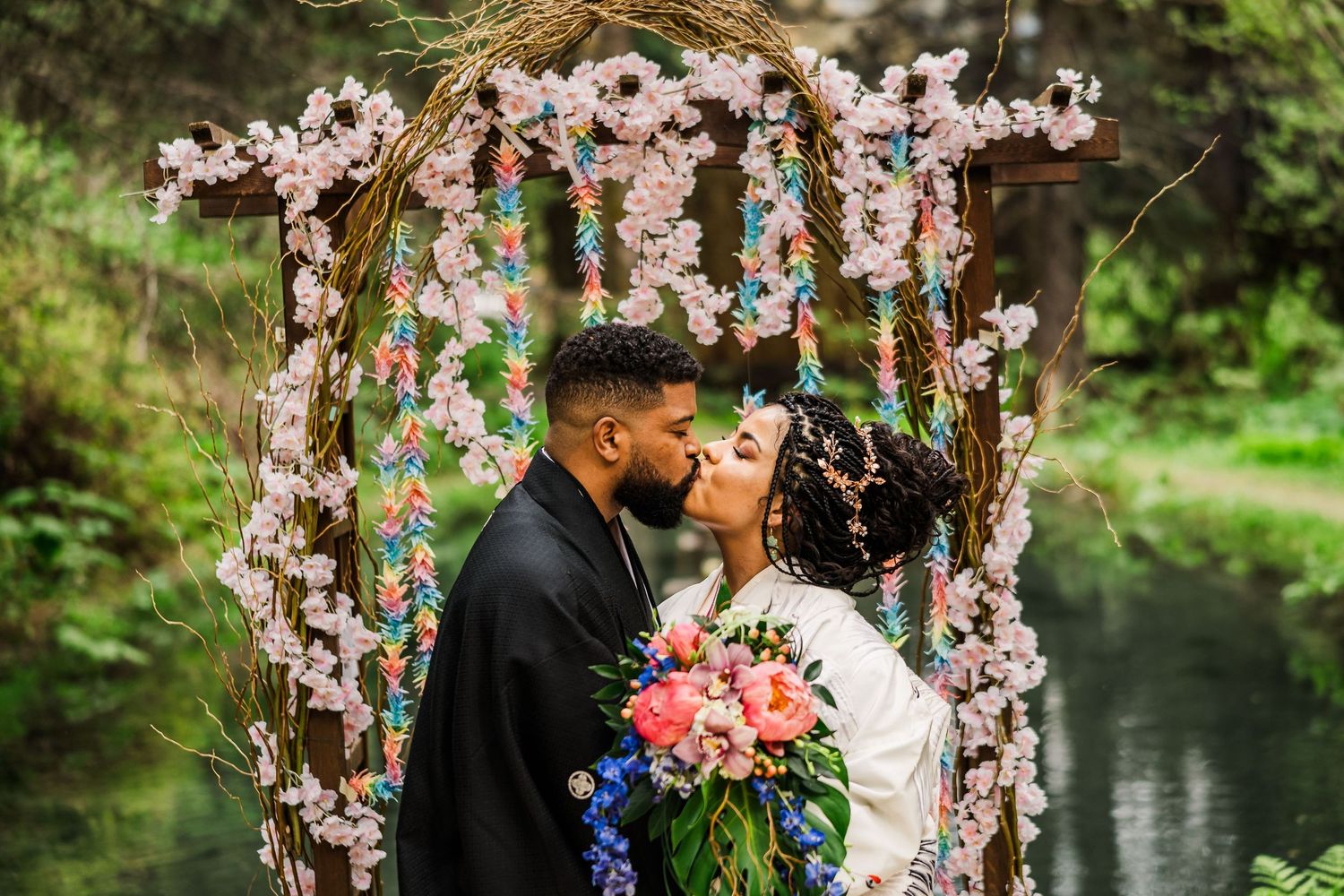 Wedding arch decorated with cherry blossoms and colorful flowers frames couple in romantic forest setting.