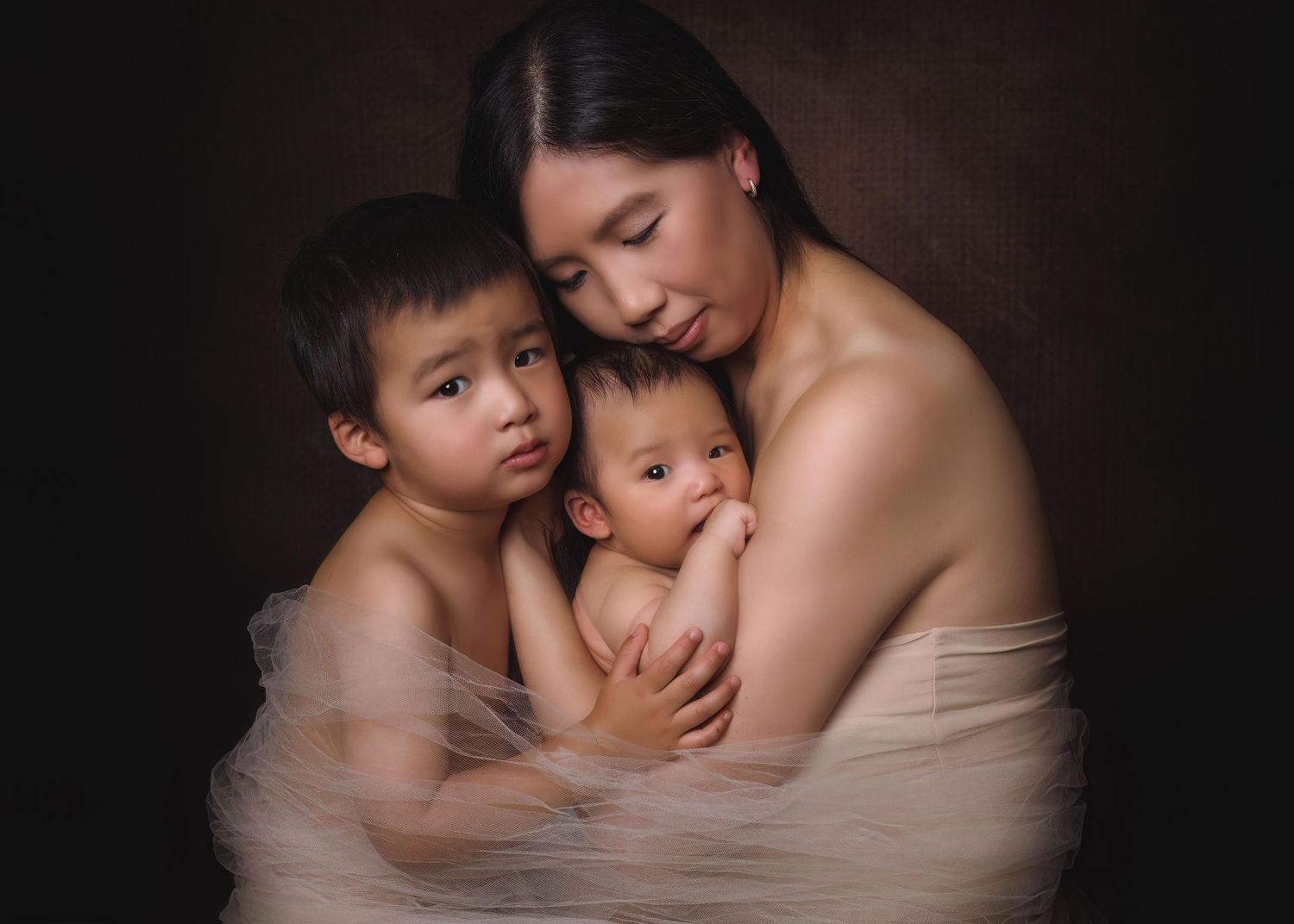 Intimate studio portrait of mother with two children embraced in soft sheer fabric against dark background.