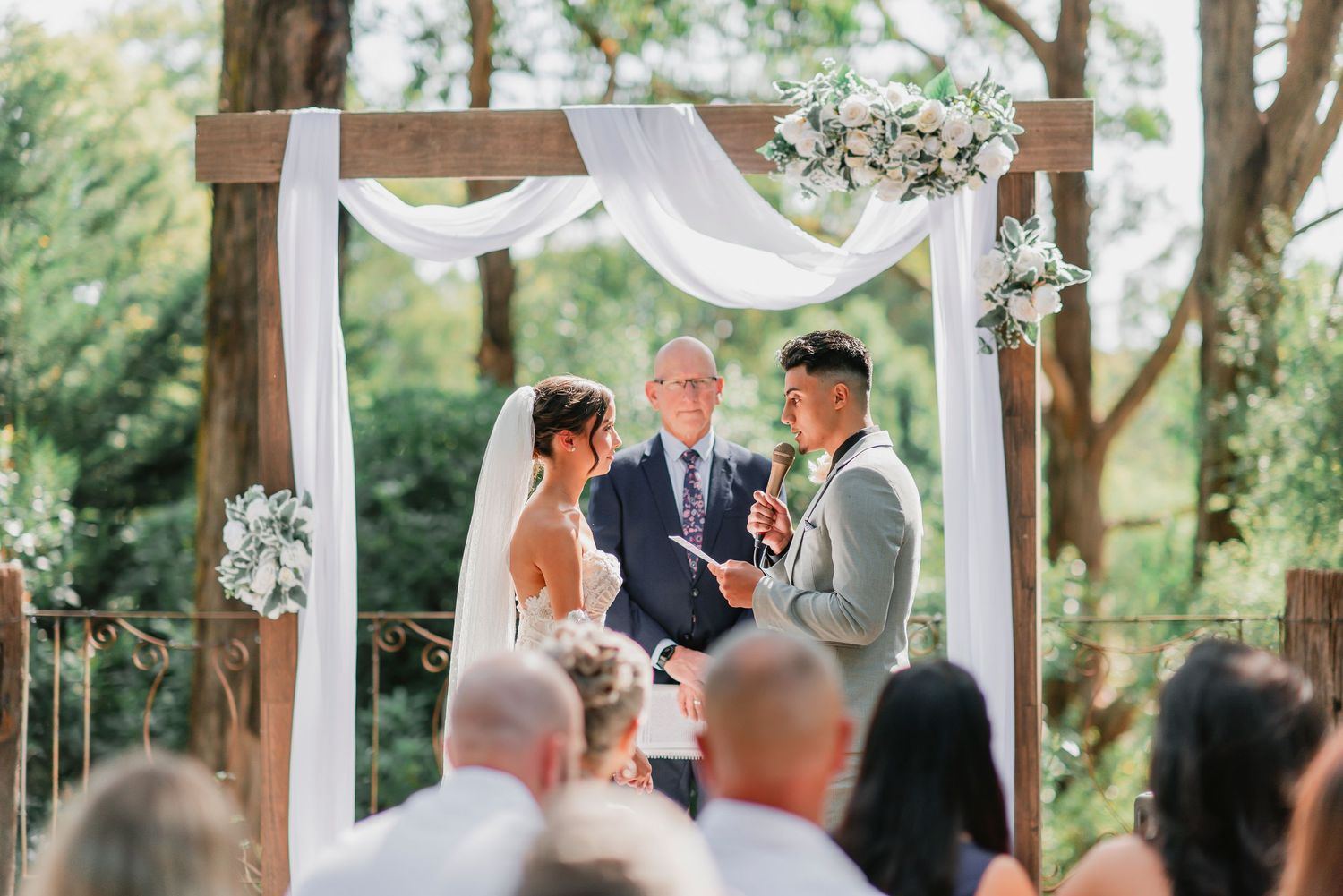 A wedding ceremony takes place under a wooden arch draped with white fabric and floral decorations in an outdoor garden setting.