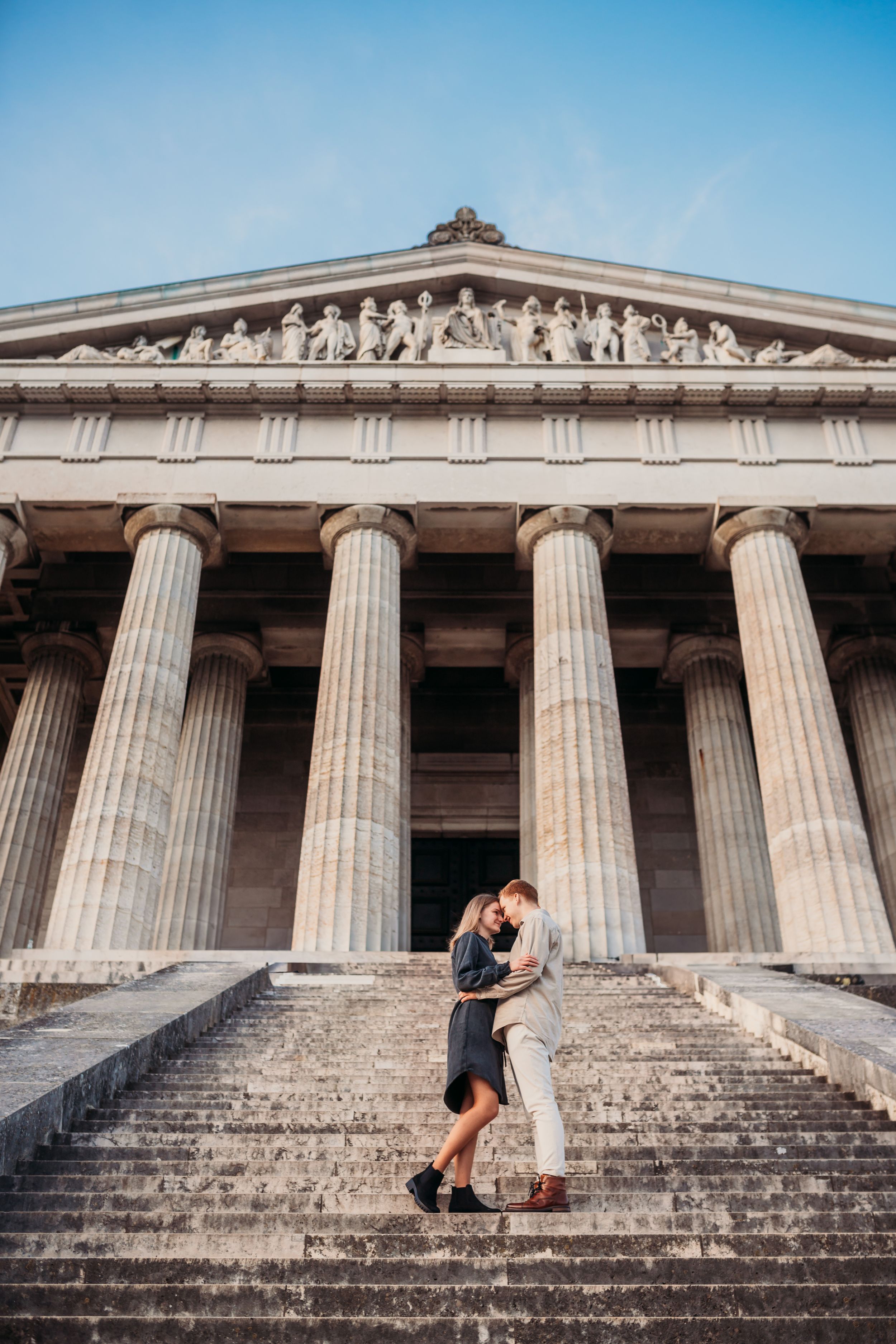 Couple embracing on steps of neoclassical building with grand columns and sculptural pediment.