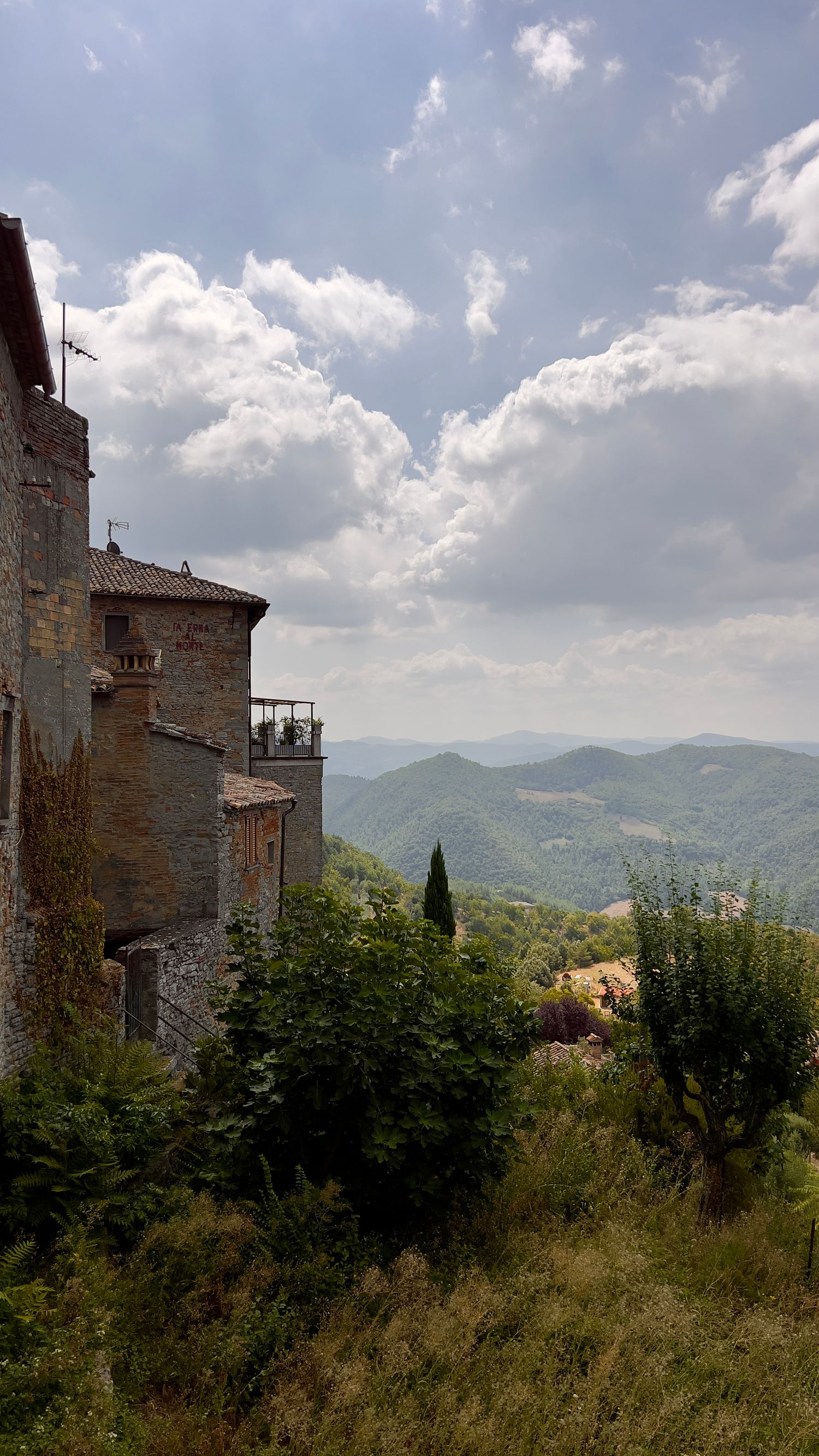 A rustic stone building perches on a hillside with rolling mountains and cypress trees in the distance.