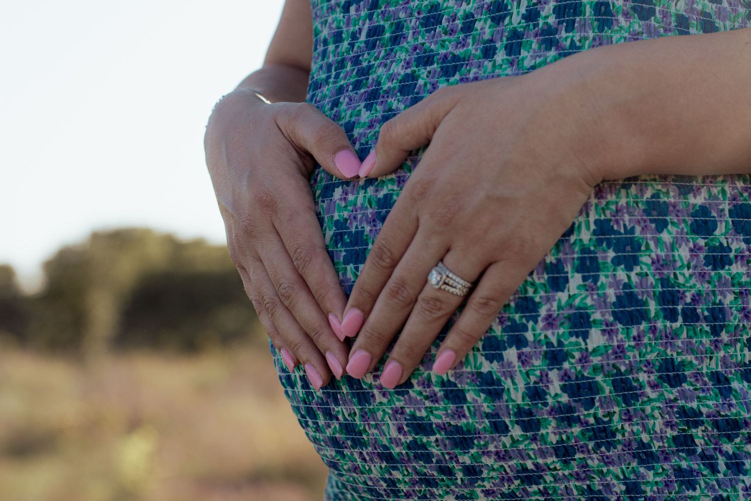 A close-up of hands resting on a pregnant belly wearing a blue and green patterned dress outdoors.