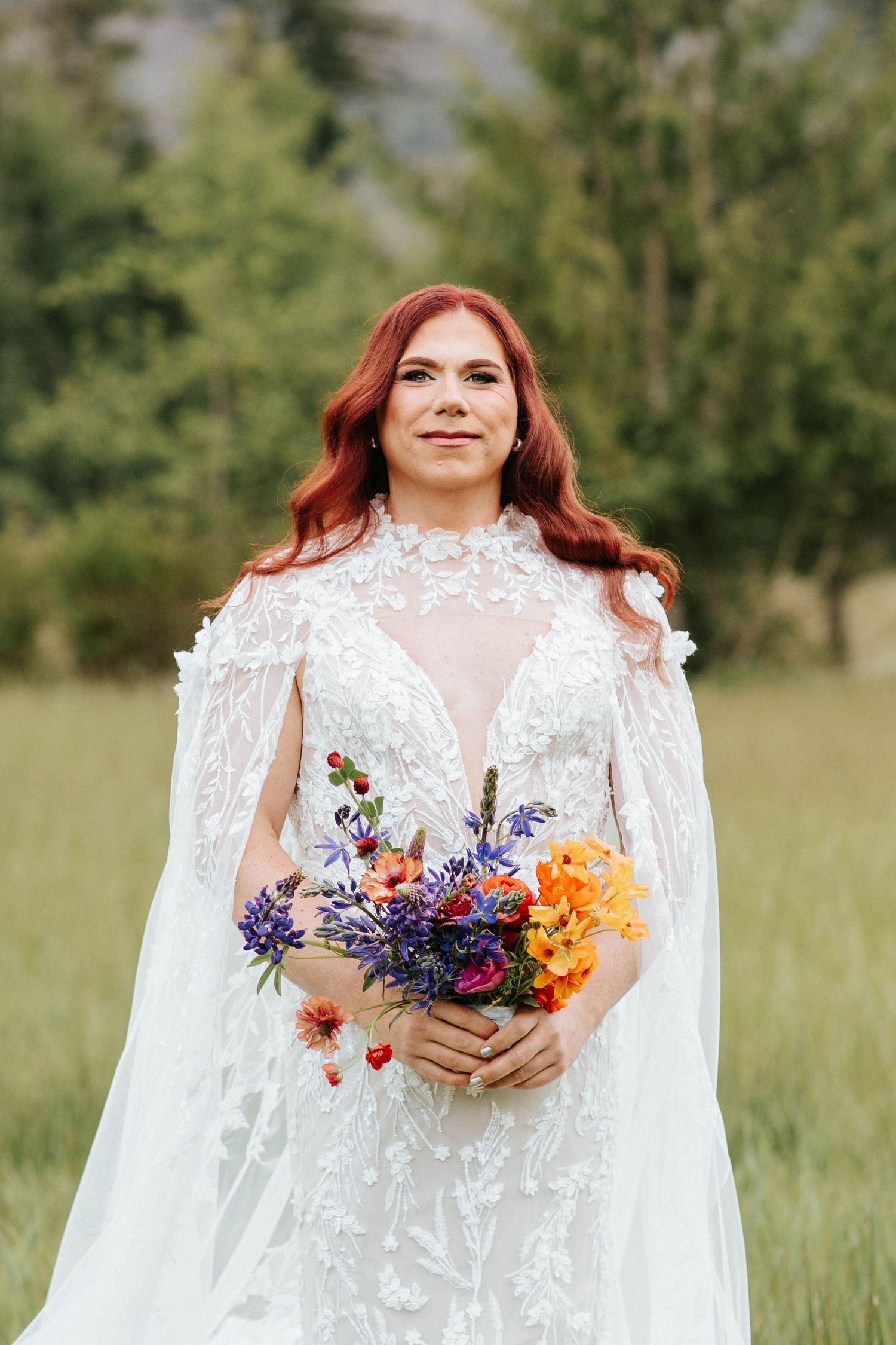 A bride in a white lace cape dress holds a vibrant bouquet of wildflowers in a sunny meadow.