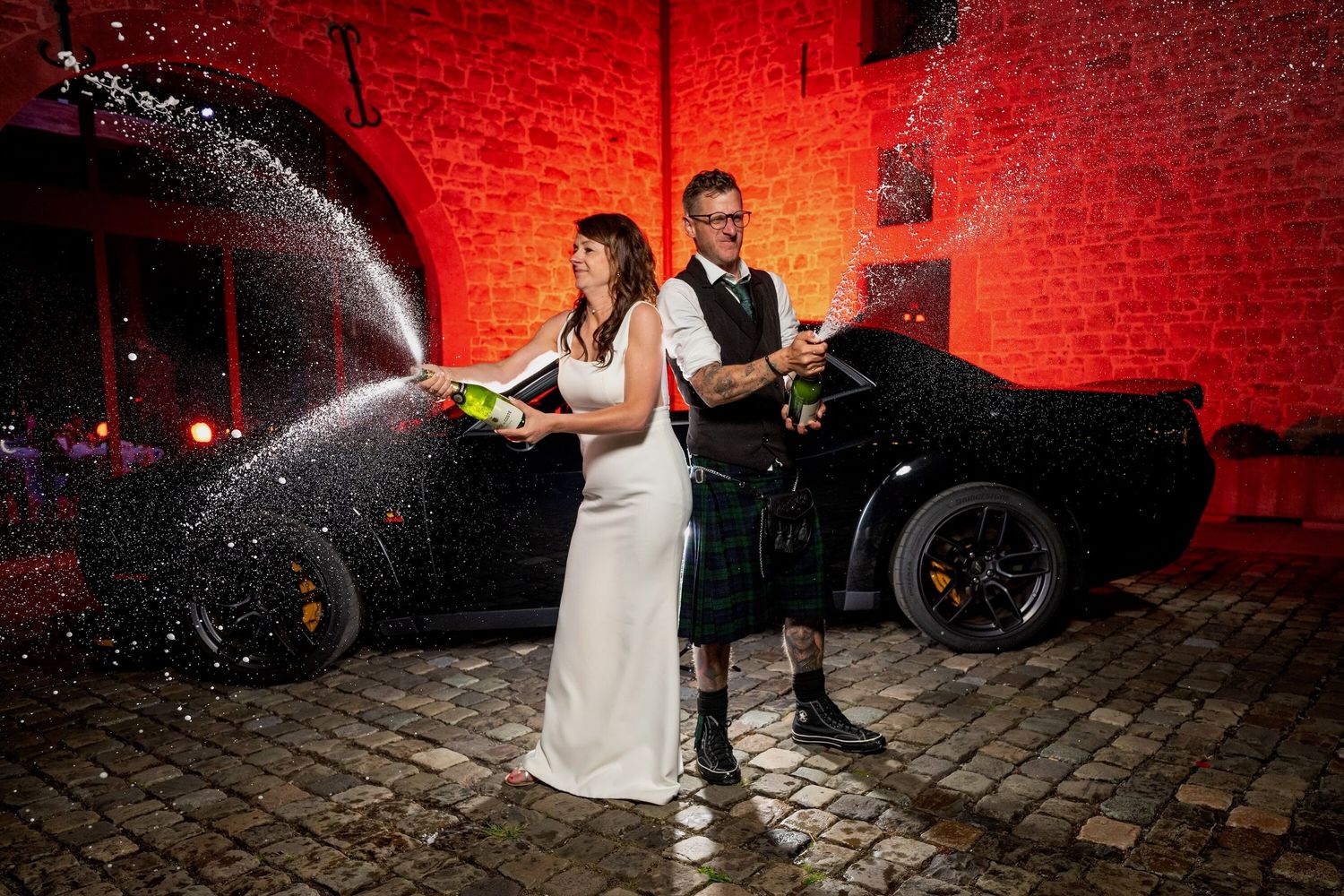 A couple in wedding attire playfully spray champagne in front of a luxury car with red-lit brick walls behind them.