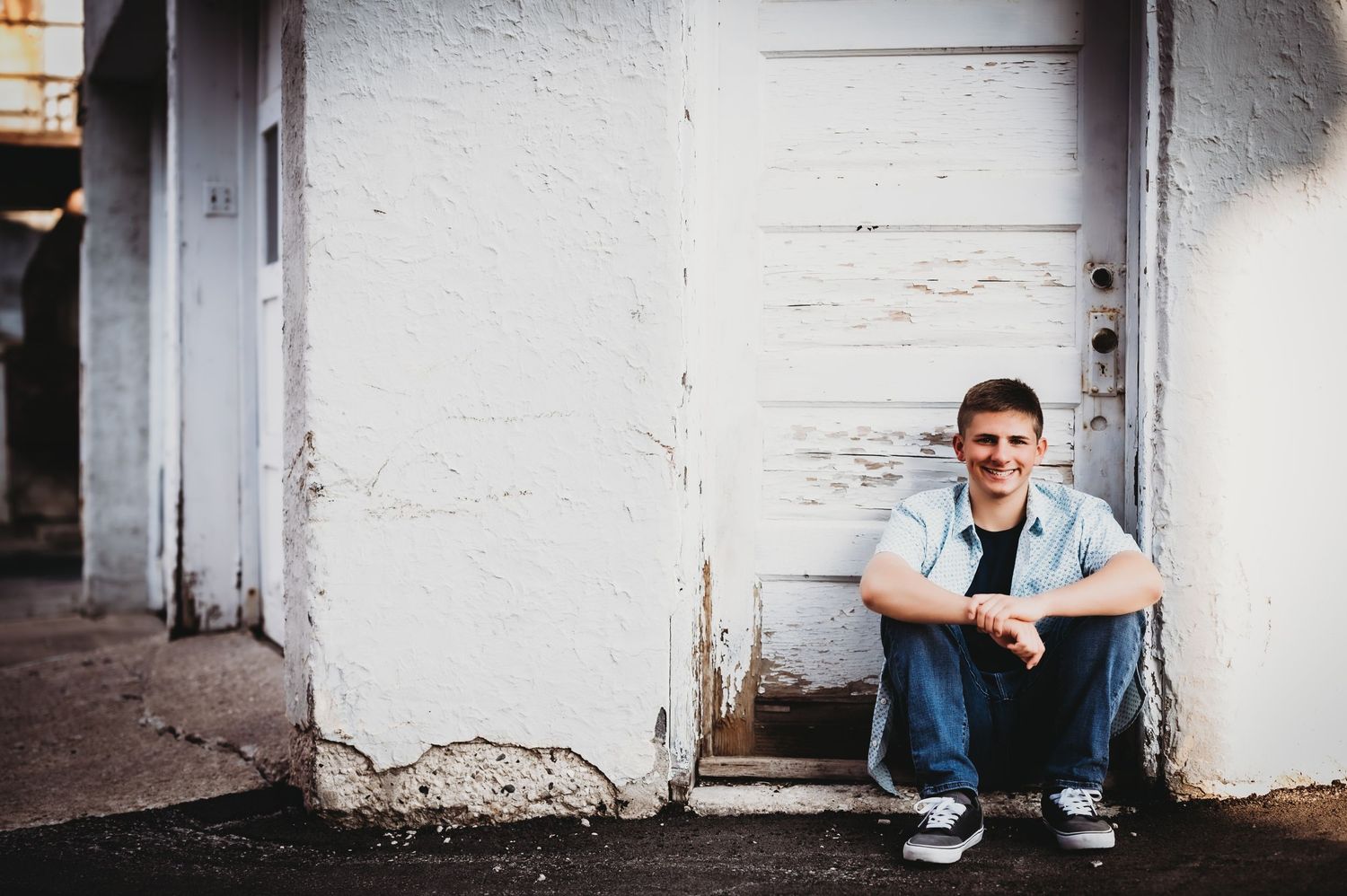 A casual portrait of someone sitting against a white wall in an urban setting.