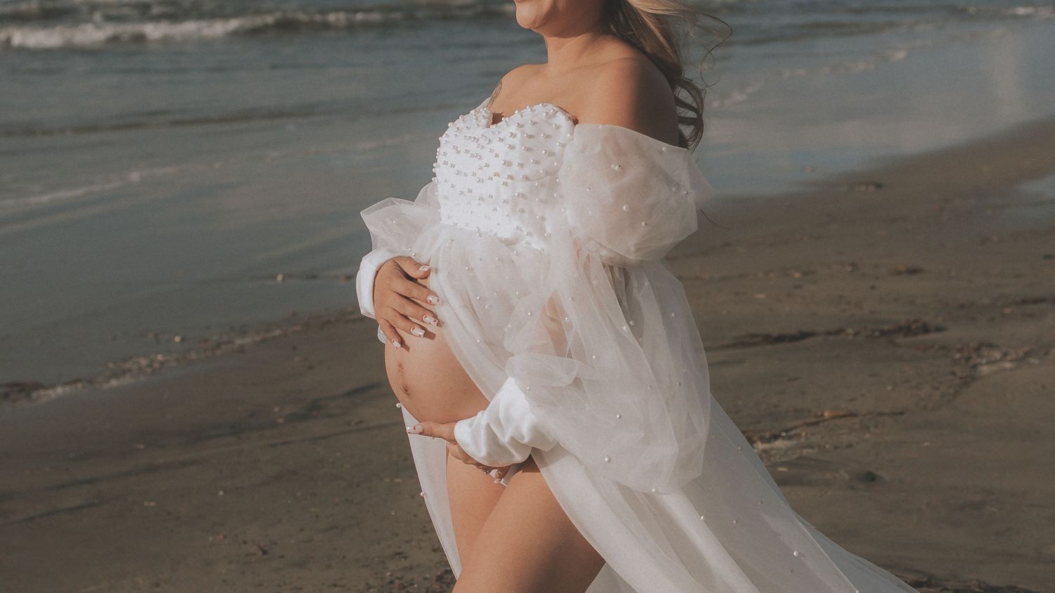 A maternity photoshoot featuring a flowing white dress on a beach at sunset.
