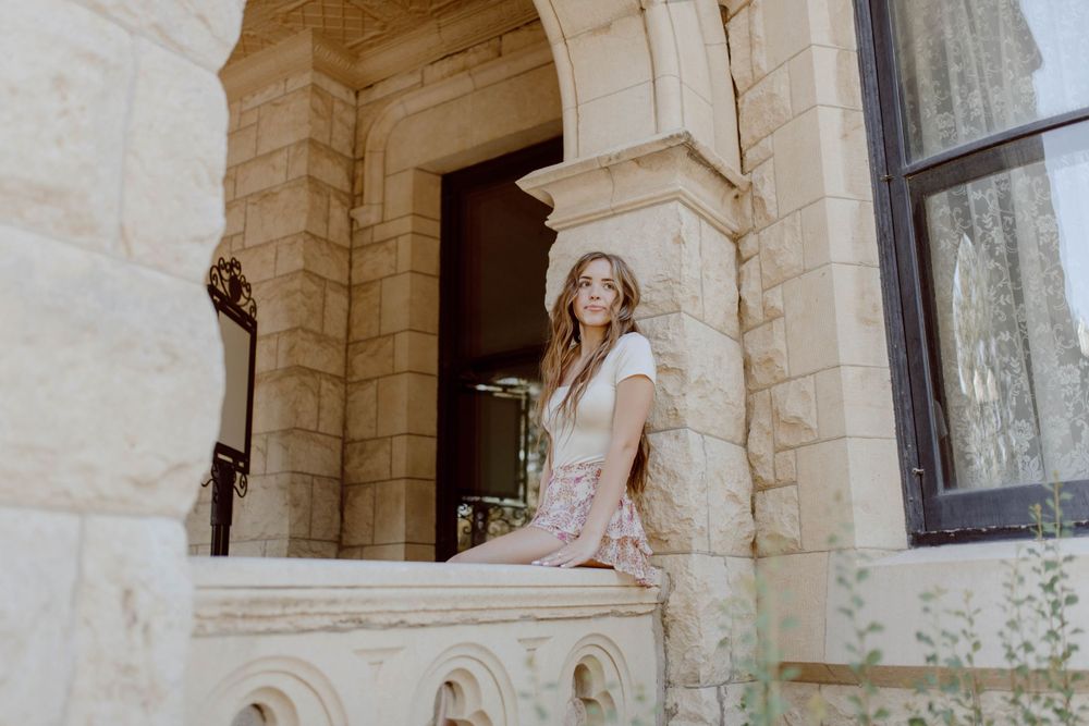 Woman in bridal gown poses on grand stone balcony of historic mansion during sunset photoshoot.