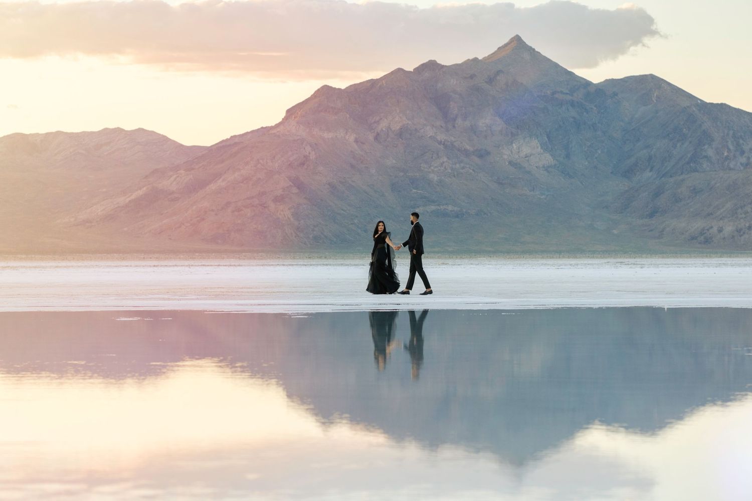 Two silhouettes stand on reflective water with misty mountains and sunset sky in the background.