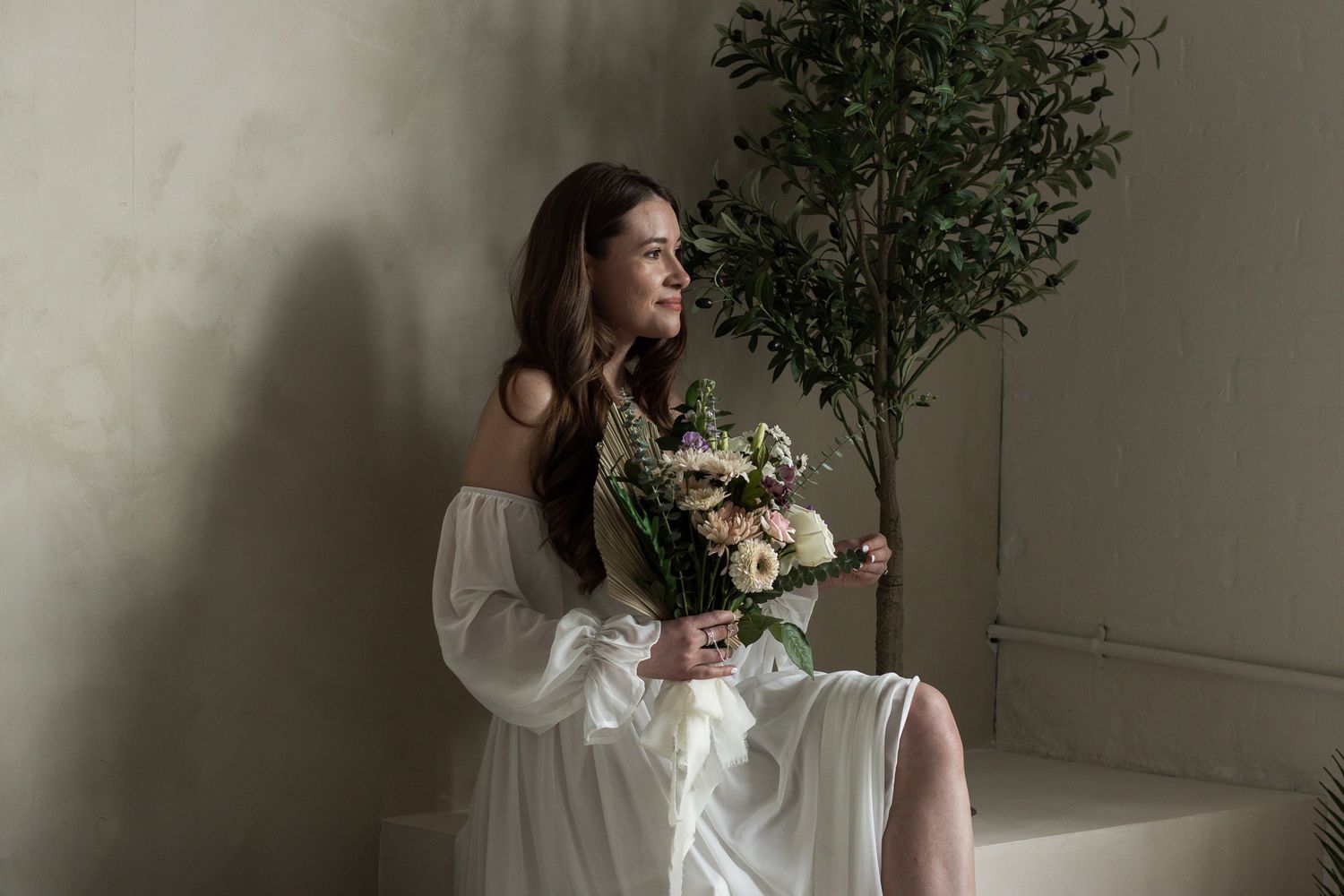 A woman in an off-shoulder white dress sits on a white surface, holding a bouquet of flowers. She is smiling and looking