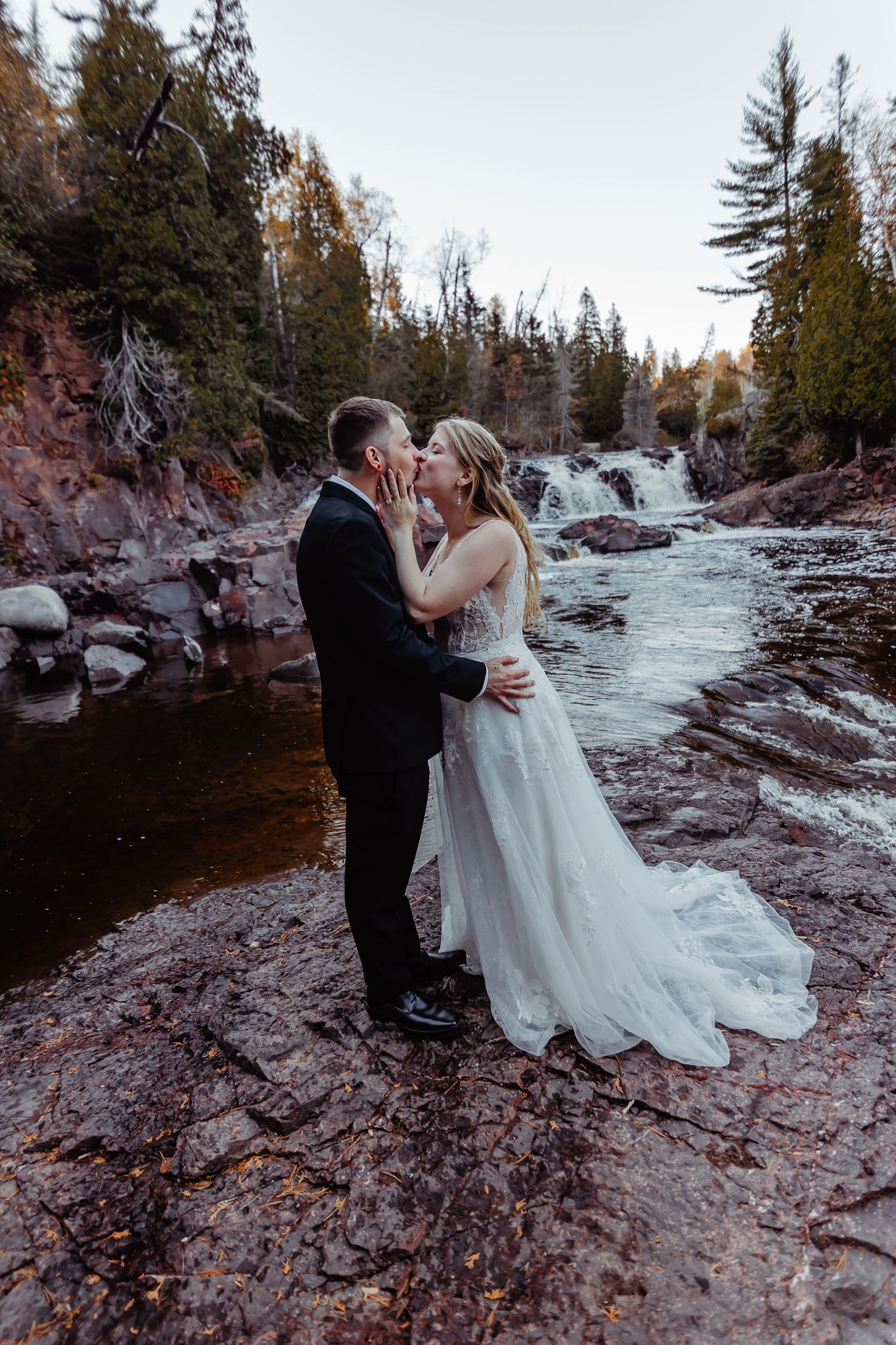 Newlyweds sharing romantic moment by waterfall with autumn trees in background.