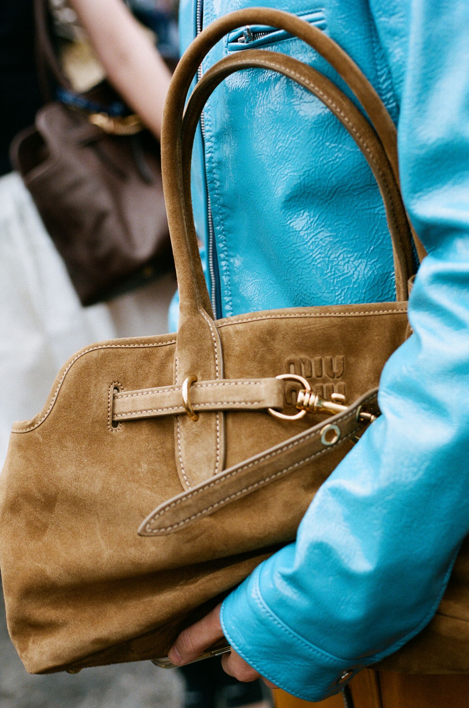 A tan suede handbag with leather straps and metal hardware being held against a turquoise blue background.