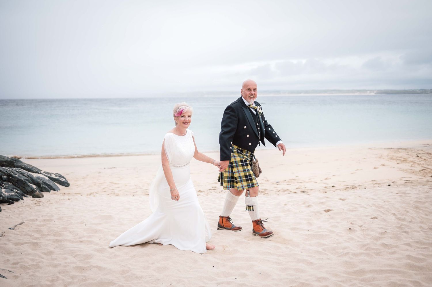 A couple walks hand in hand on a beach, with one wearing traditional Scottish kilt attire and the other in a white gown.