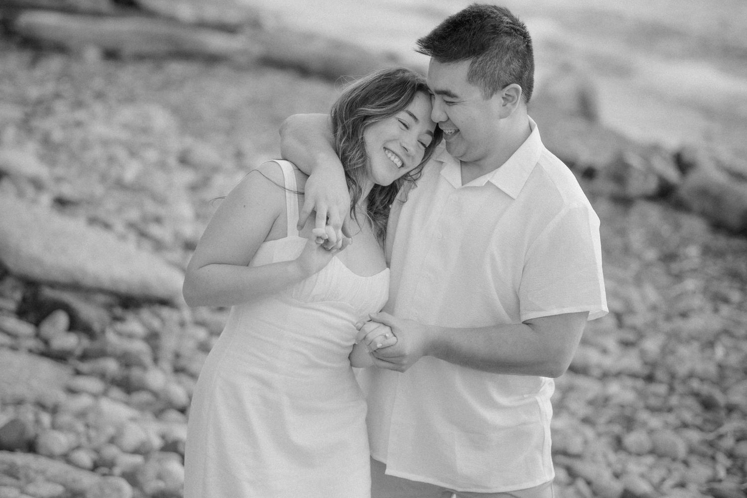 A couple laughs together during their black and white engagement photo session on a rocky shore.
