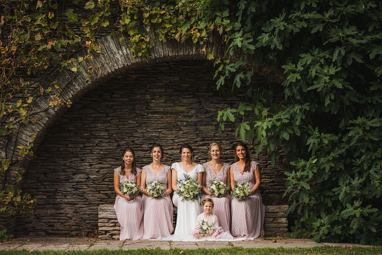 Wedding party stands under stone archway surrounded by lush greenery holding elegant floral bouquets.