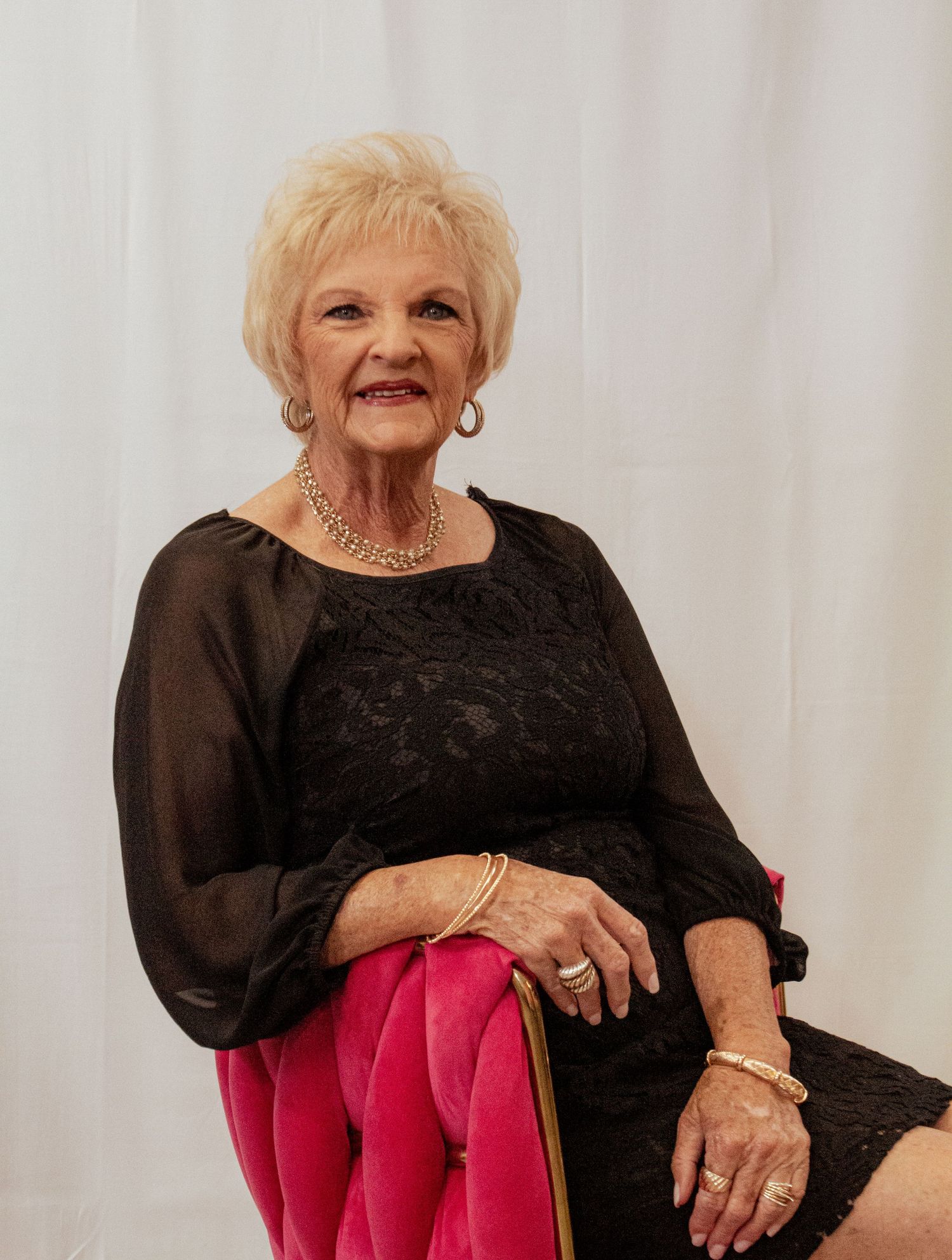 Elegant senior woman wearing black dress and pearls poses in front of white backdrop.
