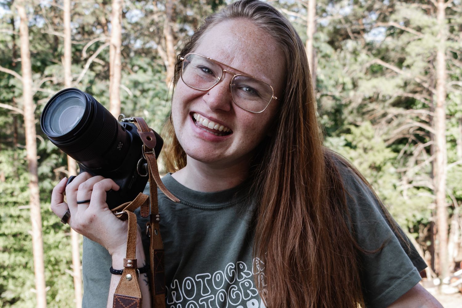 Smiling woman with long red hair and glasses holding a camera, standing outdoors with trees in the background