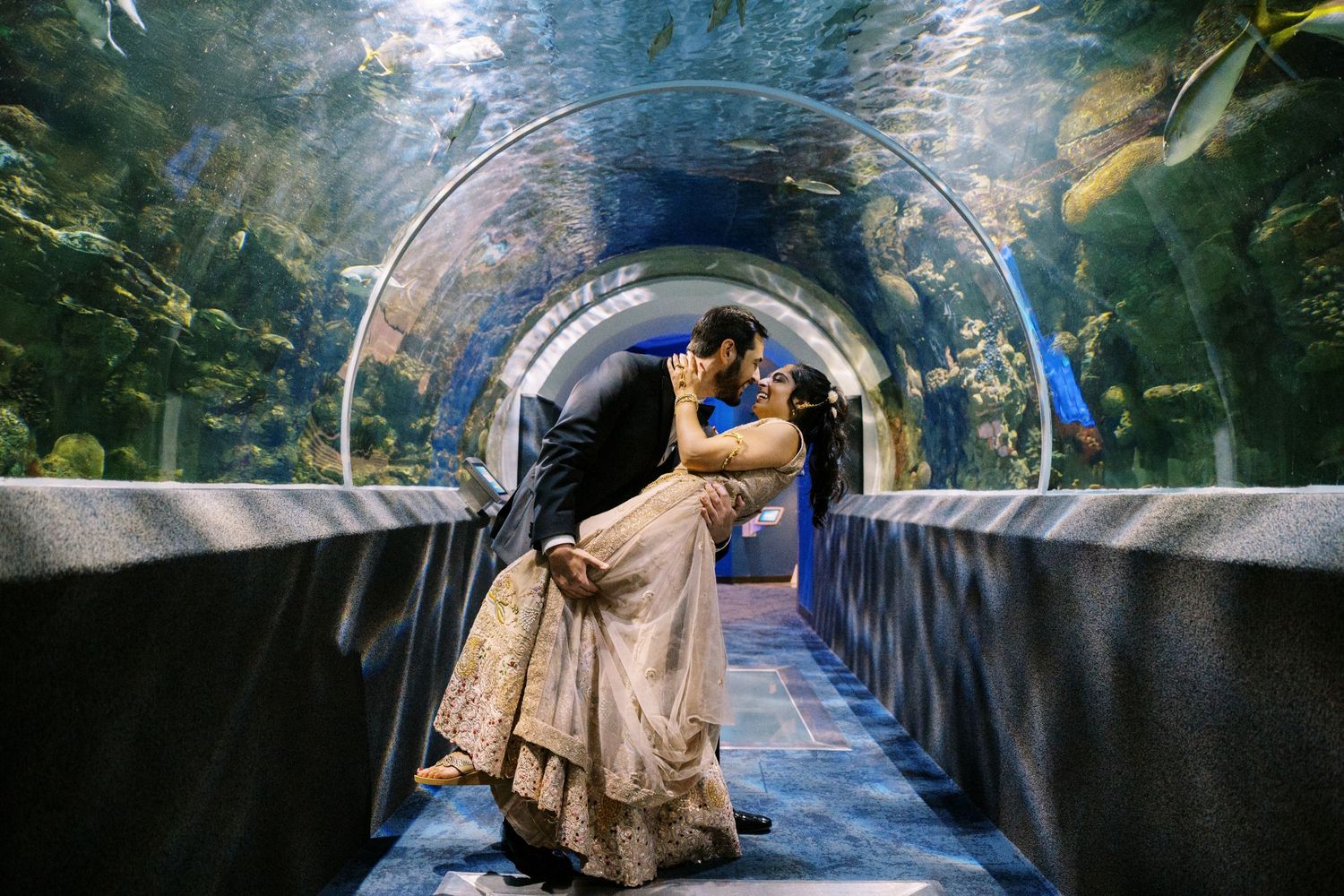 A couple shares a romantic moment in a curved aquarium tunnel walkway surrounded by blue water and marine life.