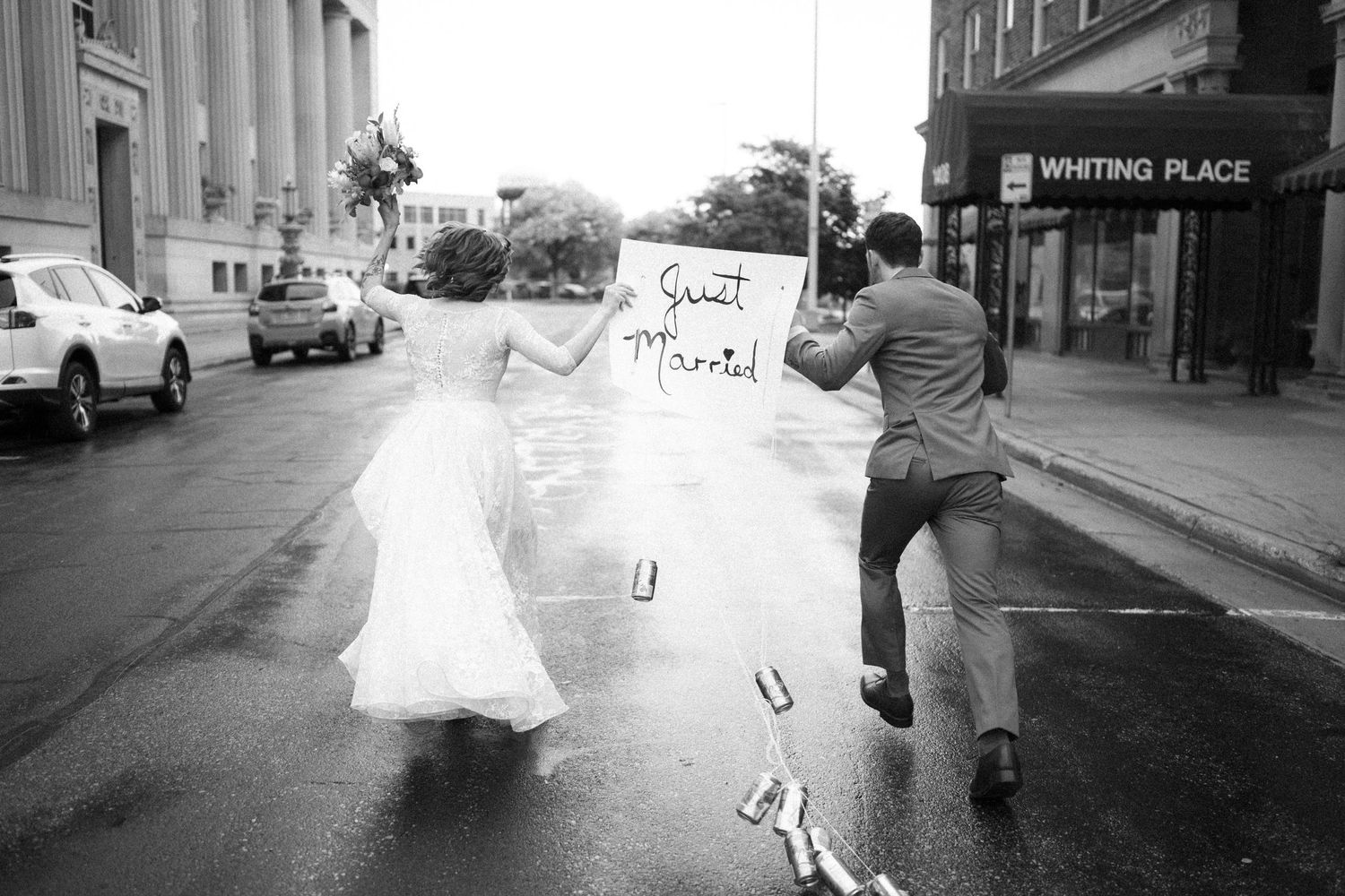 Black and white photo of newlyweds running through downtown streets as they hold a "just married" sign.