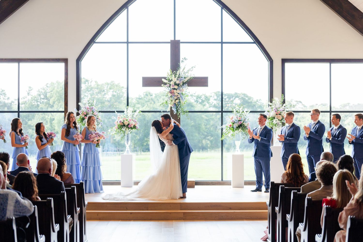 Wedding ceremony in bright chapel with large arched window, blue-clad bridesmaids and wooden pews filled with guests.