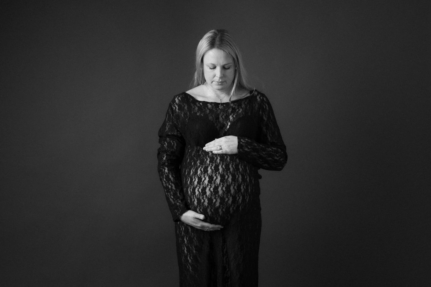 gravid kvinna i svart spetsklänning står framför en grå bakgrund vid gravidfotografering i studio i Mullsjö / Jönköping. Hon håller om magen