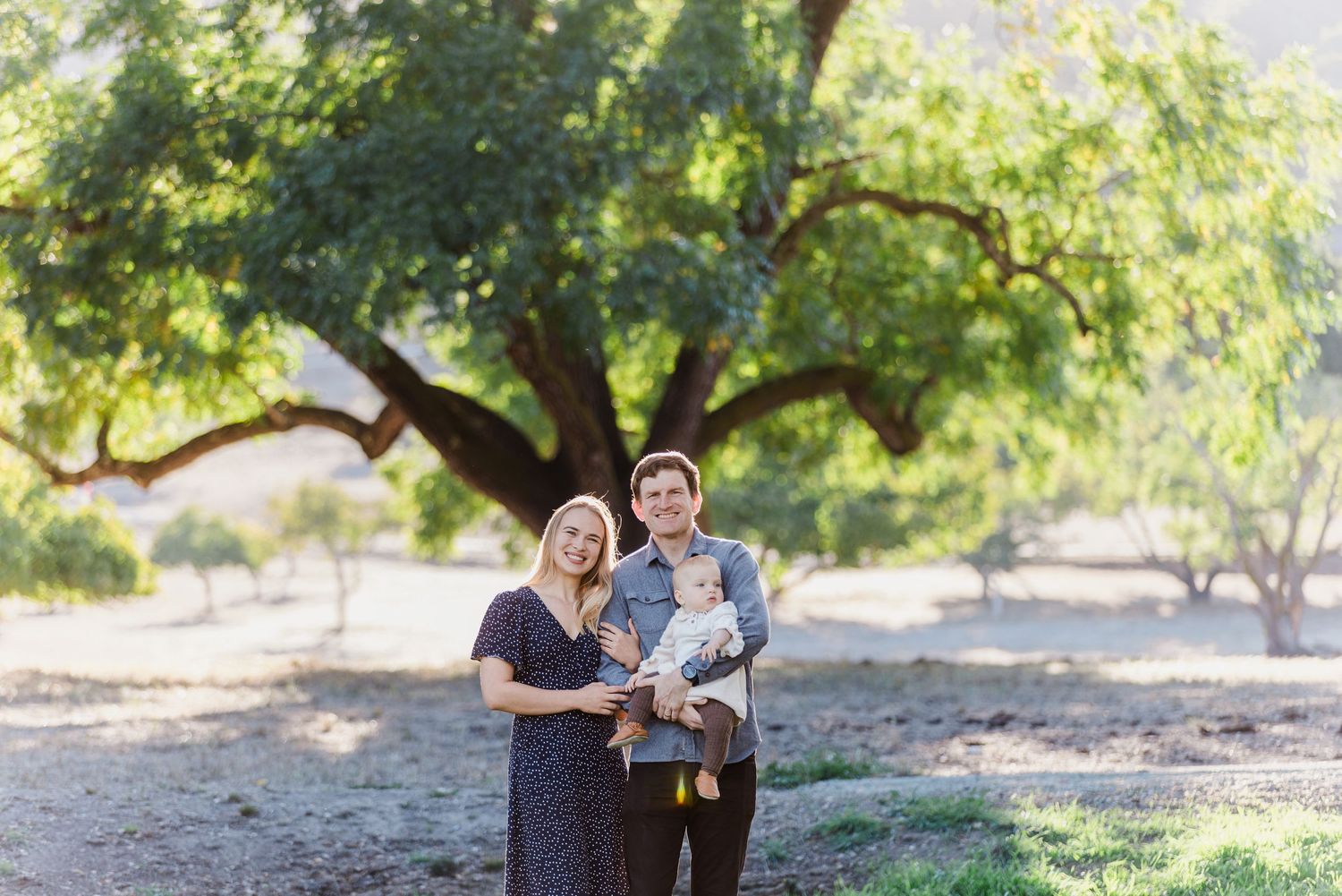 A family poses together under a large, sprawling tree on a sunny day.
