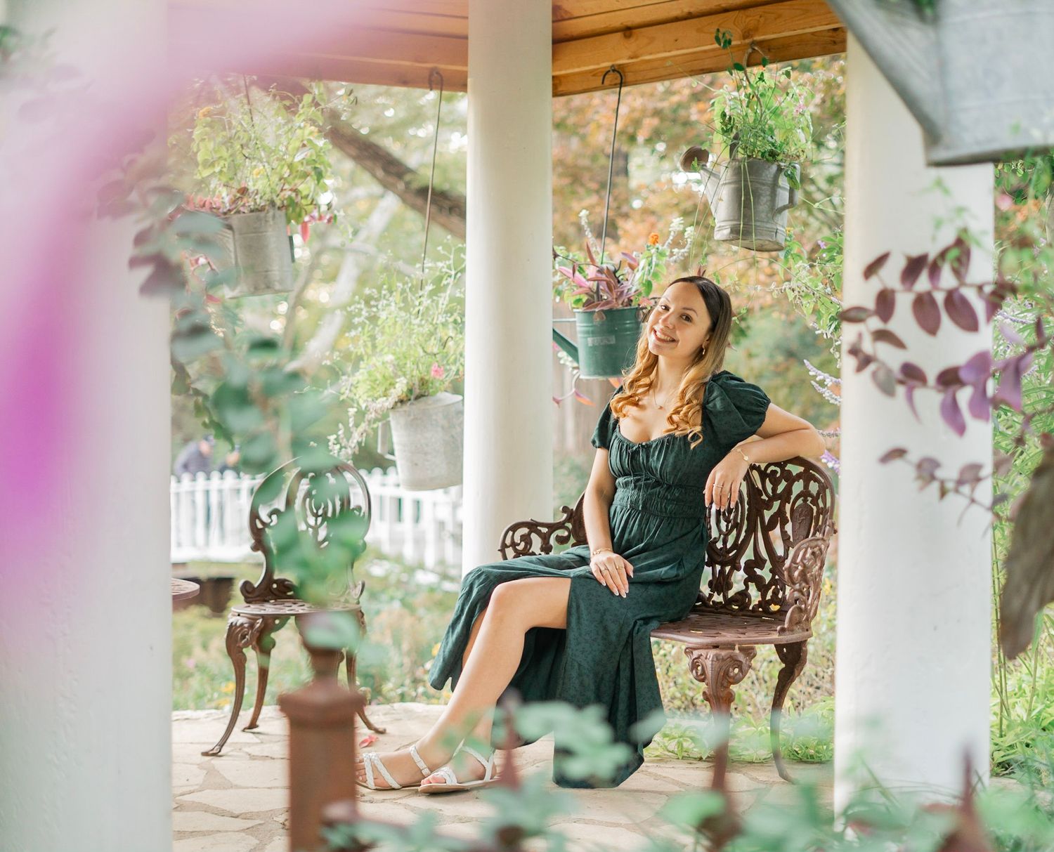 Young woman in a dark green dress sitting on ornate chair on a plant-filled porch in autumn.