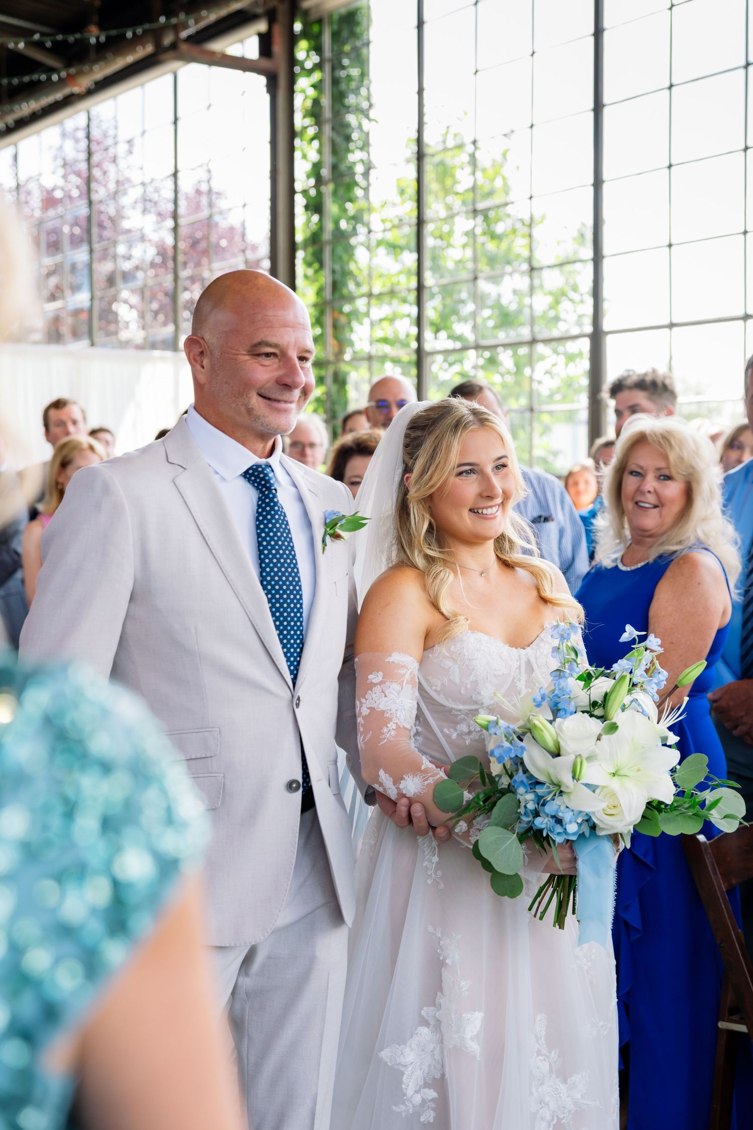 A joyful wedding celebration with the couple smiling during their ceremony, surrounded by guests and blue floral decor.