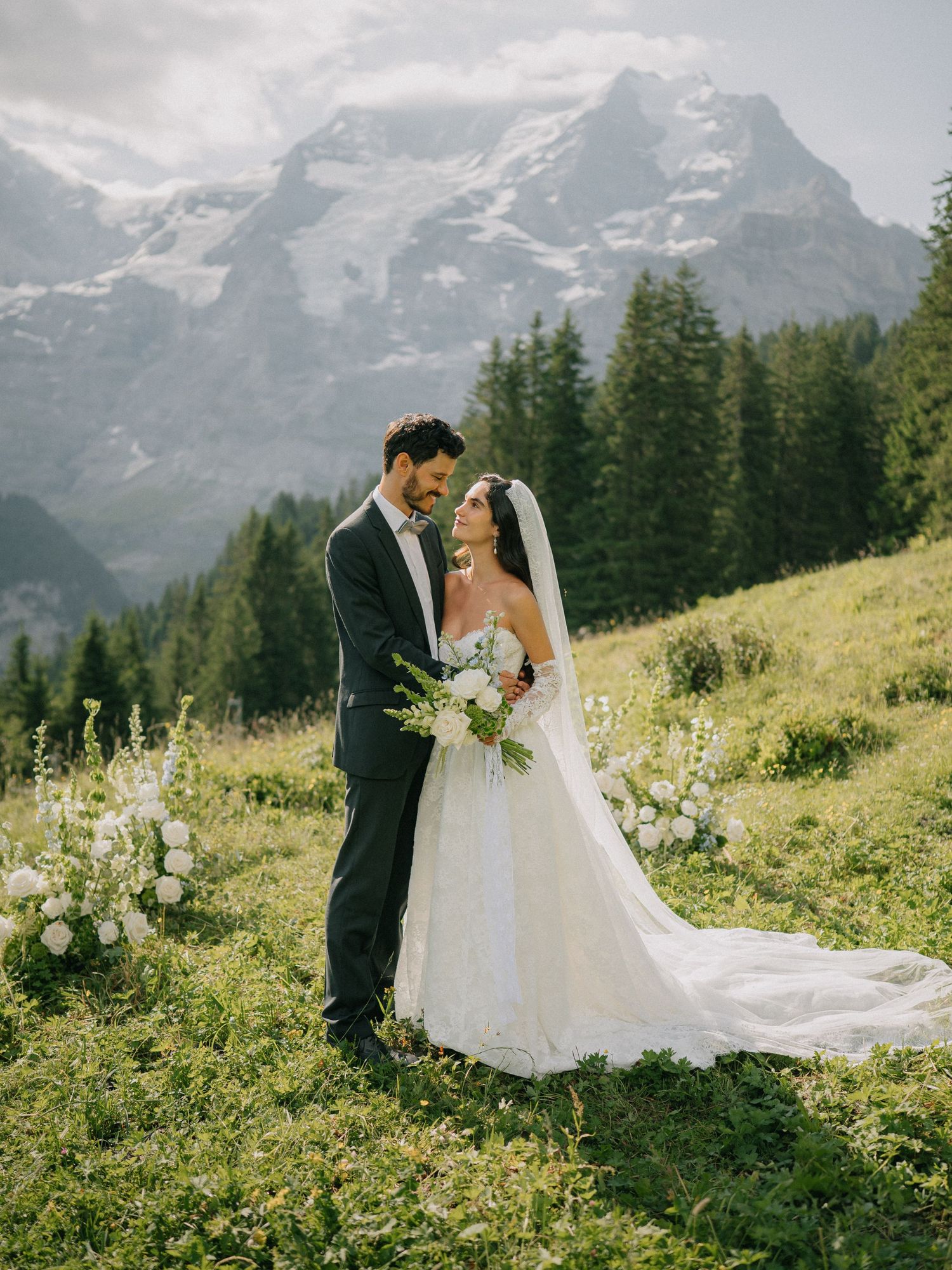 Wedding couple stands together in front of misty mountain landscape with dramatic clouds and evergreen trees.