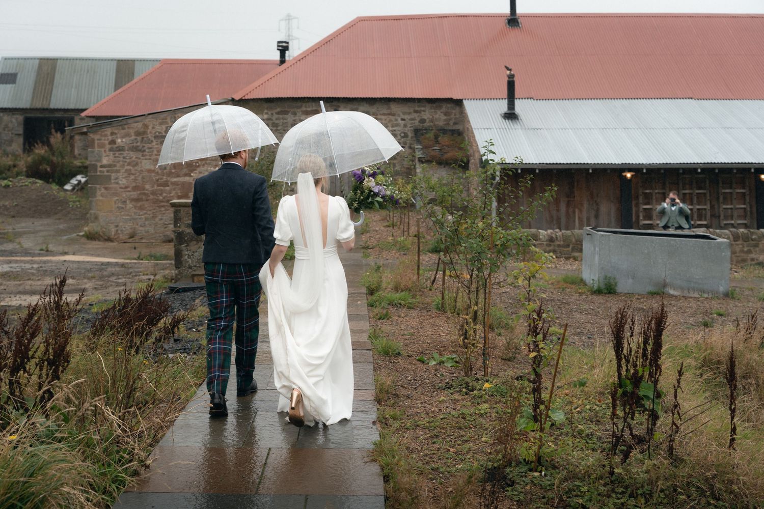 Couple walking through a garden path in wedding attire on a rainy day with umbrellas.
