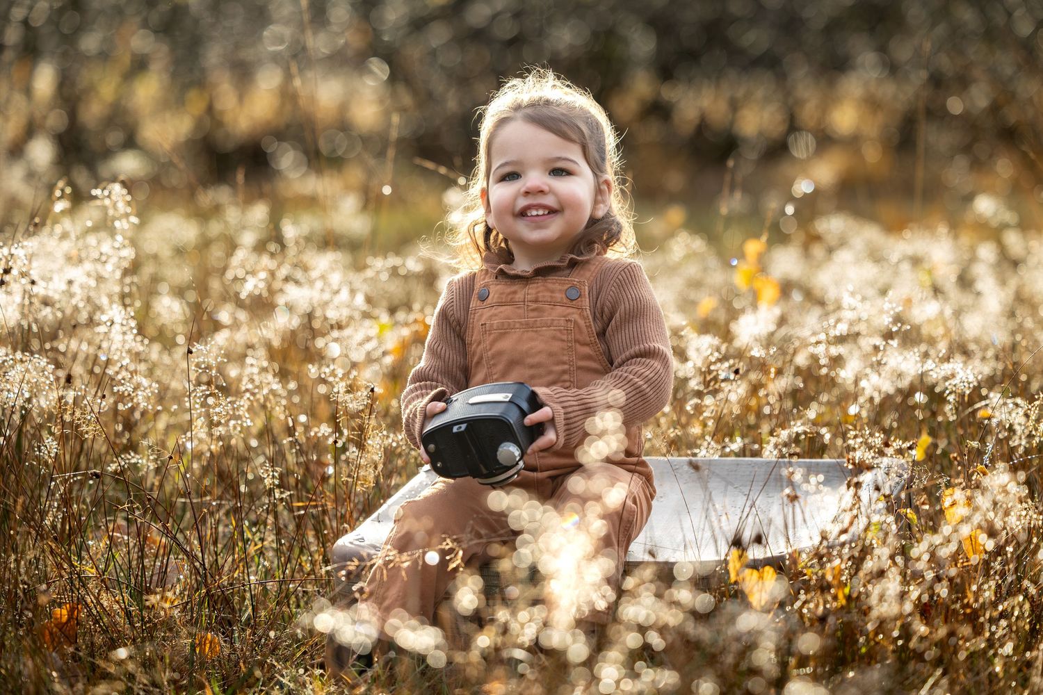 jong meisje met bruine tuinbroek, zittend op een koffer met oude camera in de hand. Tijdens een fotoshoot door Inge Muller Fotografie