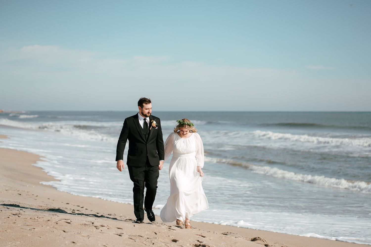 Bride and groom walking along the Carolina beach shoreline with ocean waves crashing in the background on their wedding day. North Carolina