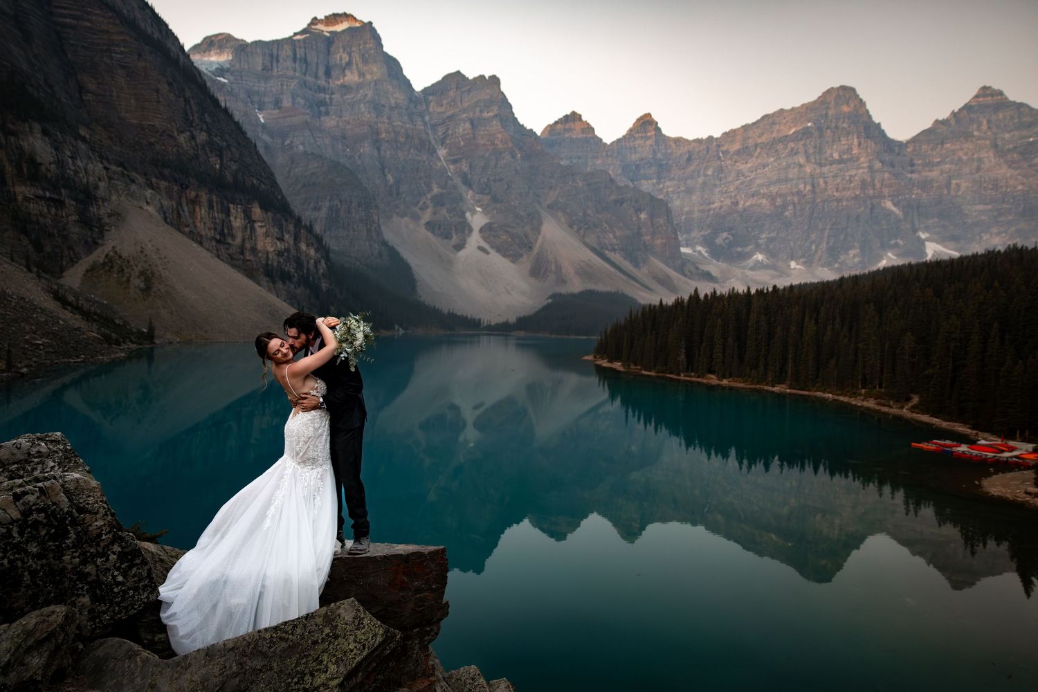 A couple embraces on a rocky overlook at Moraine Lake with the Valley of Ten Peaks reflecting in turquoise waters.