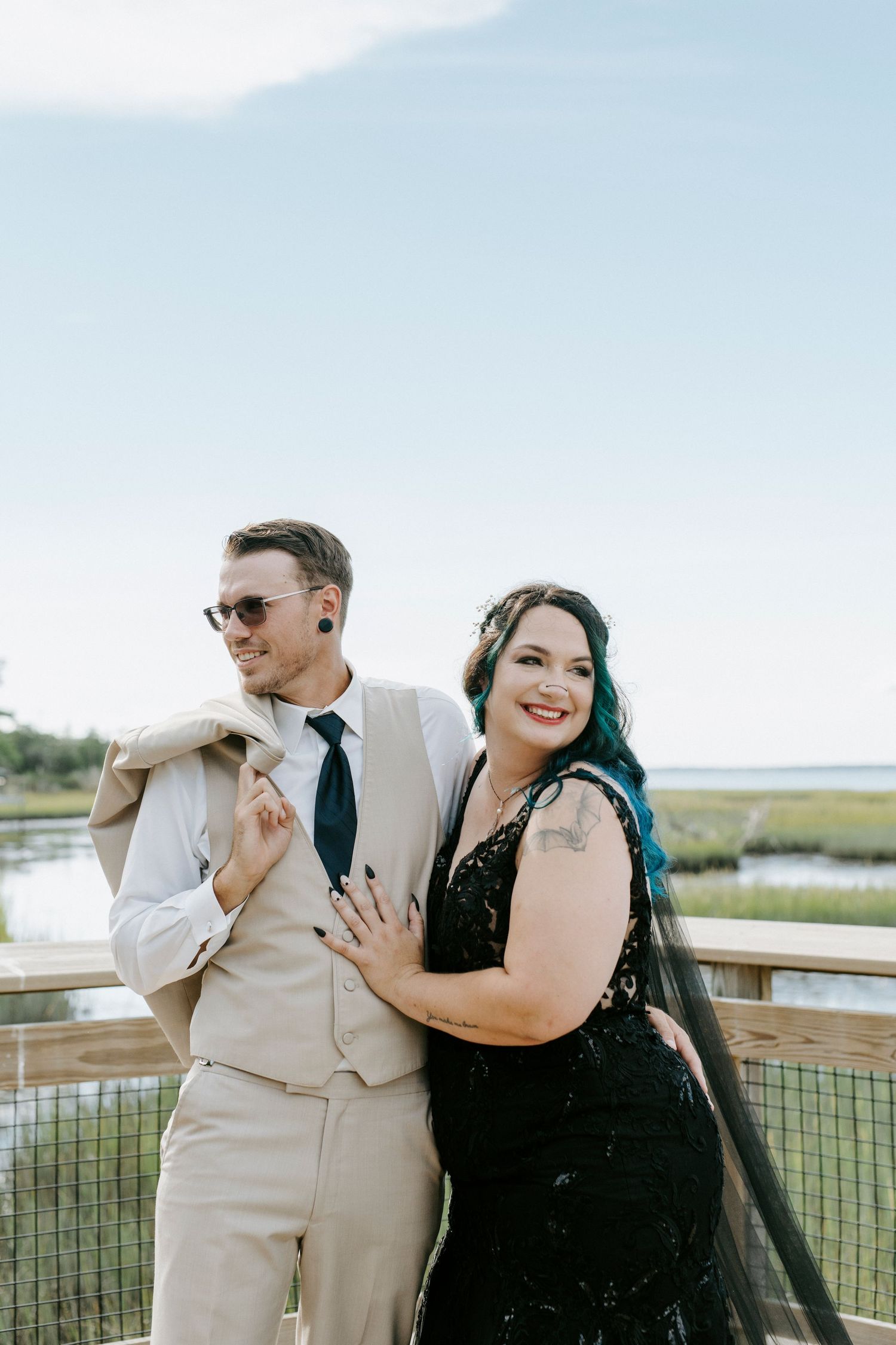 Couple in coordinated black and beige outfits share candid laughs while posing on outdoor deck by marshland.