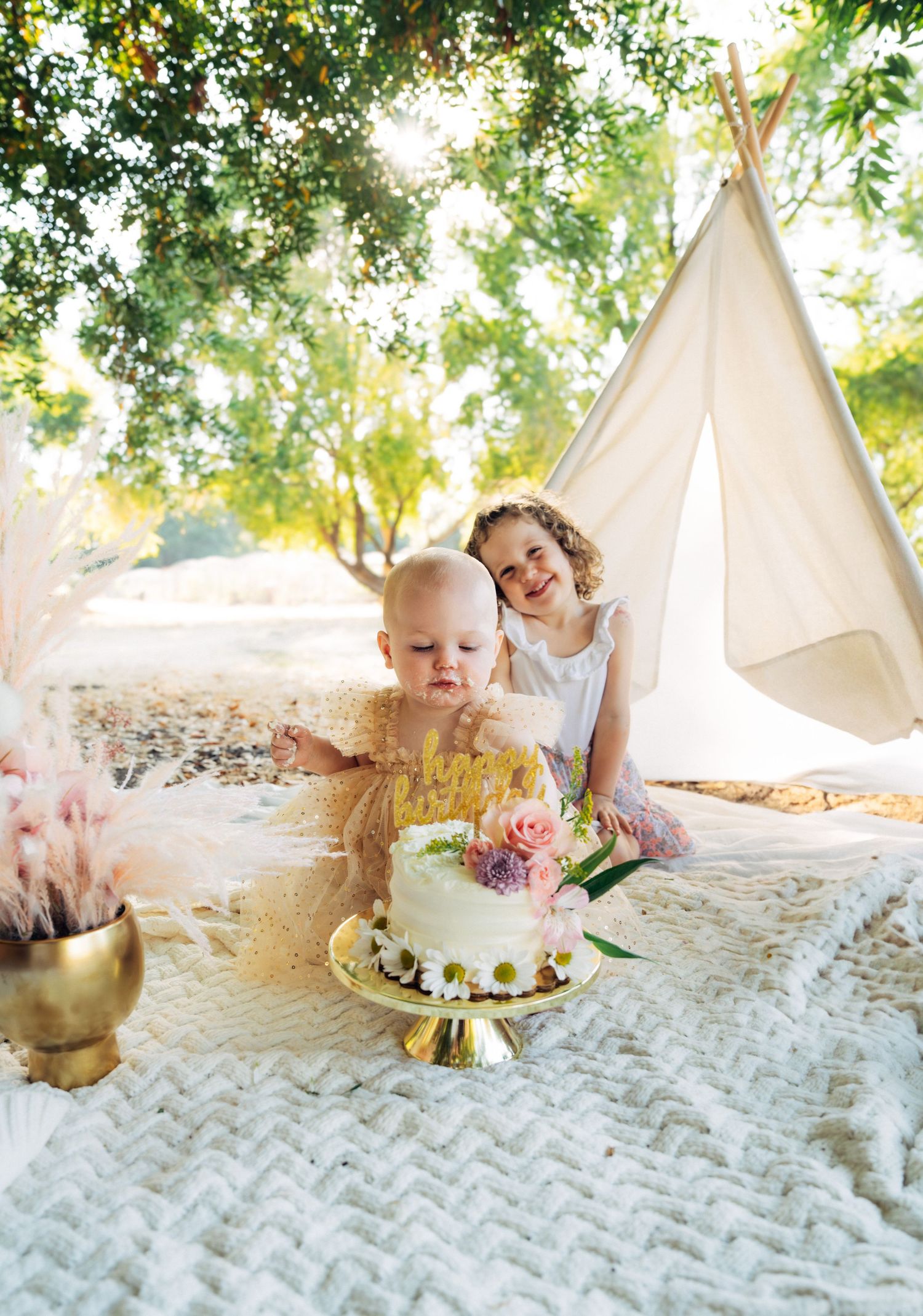 Baby celebrating first birthday milestone with cake smash photos on white blanket in outdoor garden setting.
