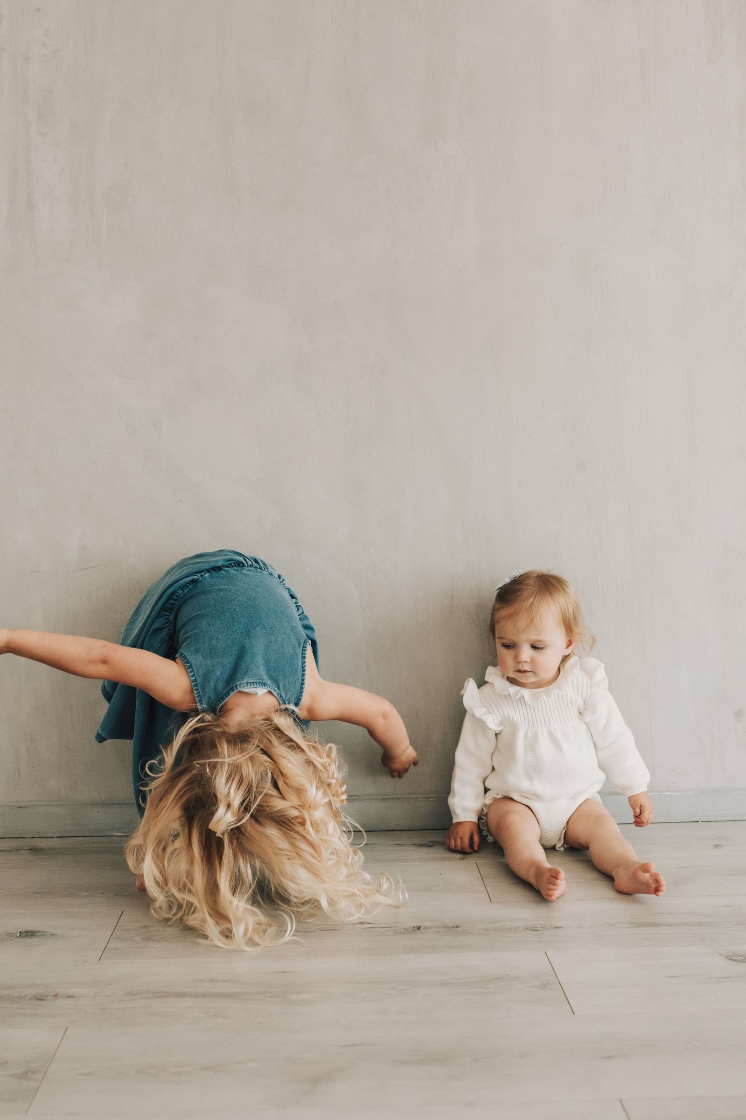 Woman in teal dress and baby lying on gray floor while doing playful rolling movements.