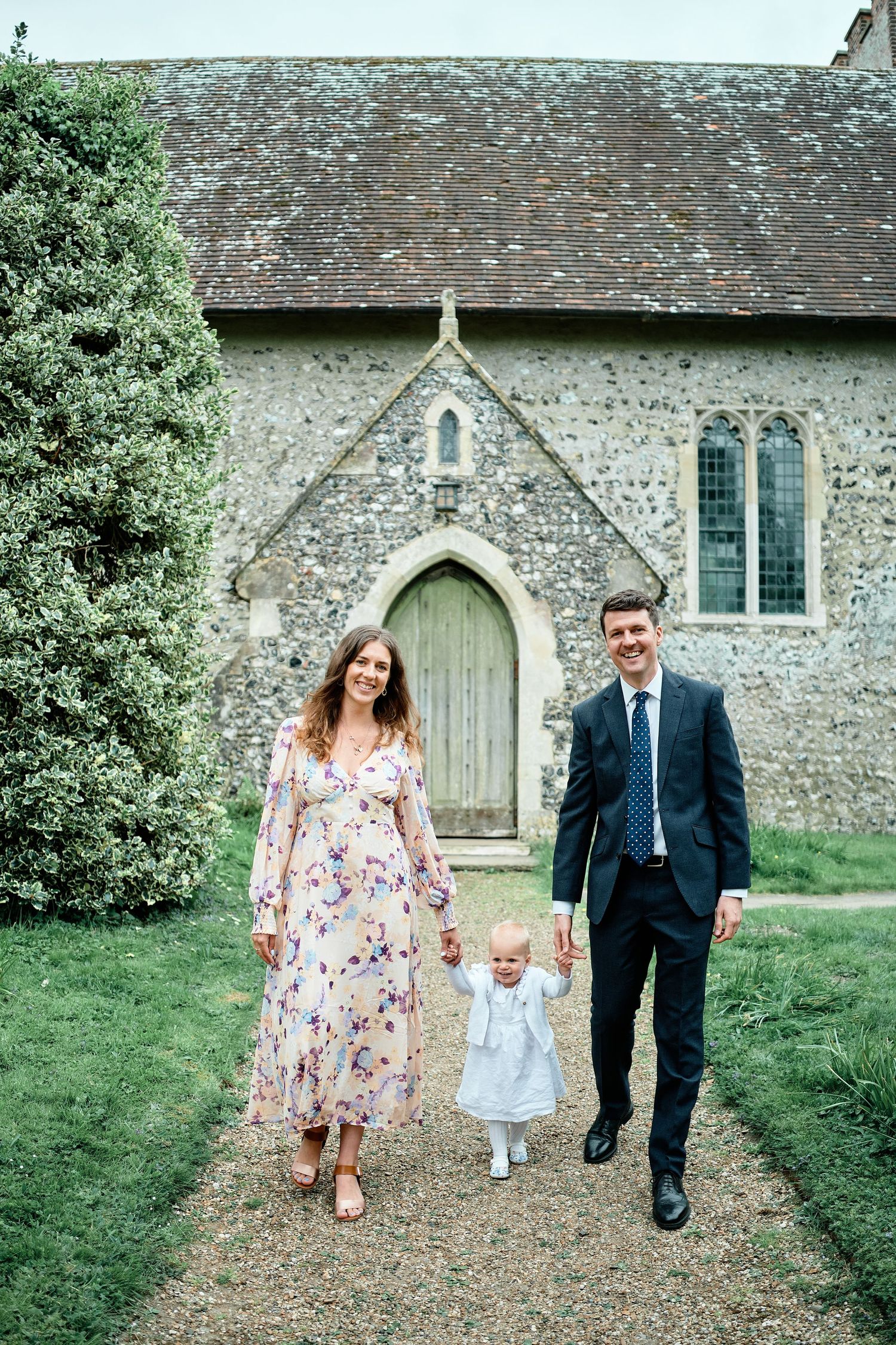 A family walks along a gravel path in front of a historic stone church with a green wooden door and stained glass windows.
