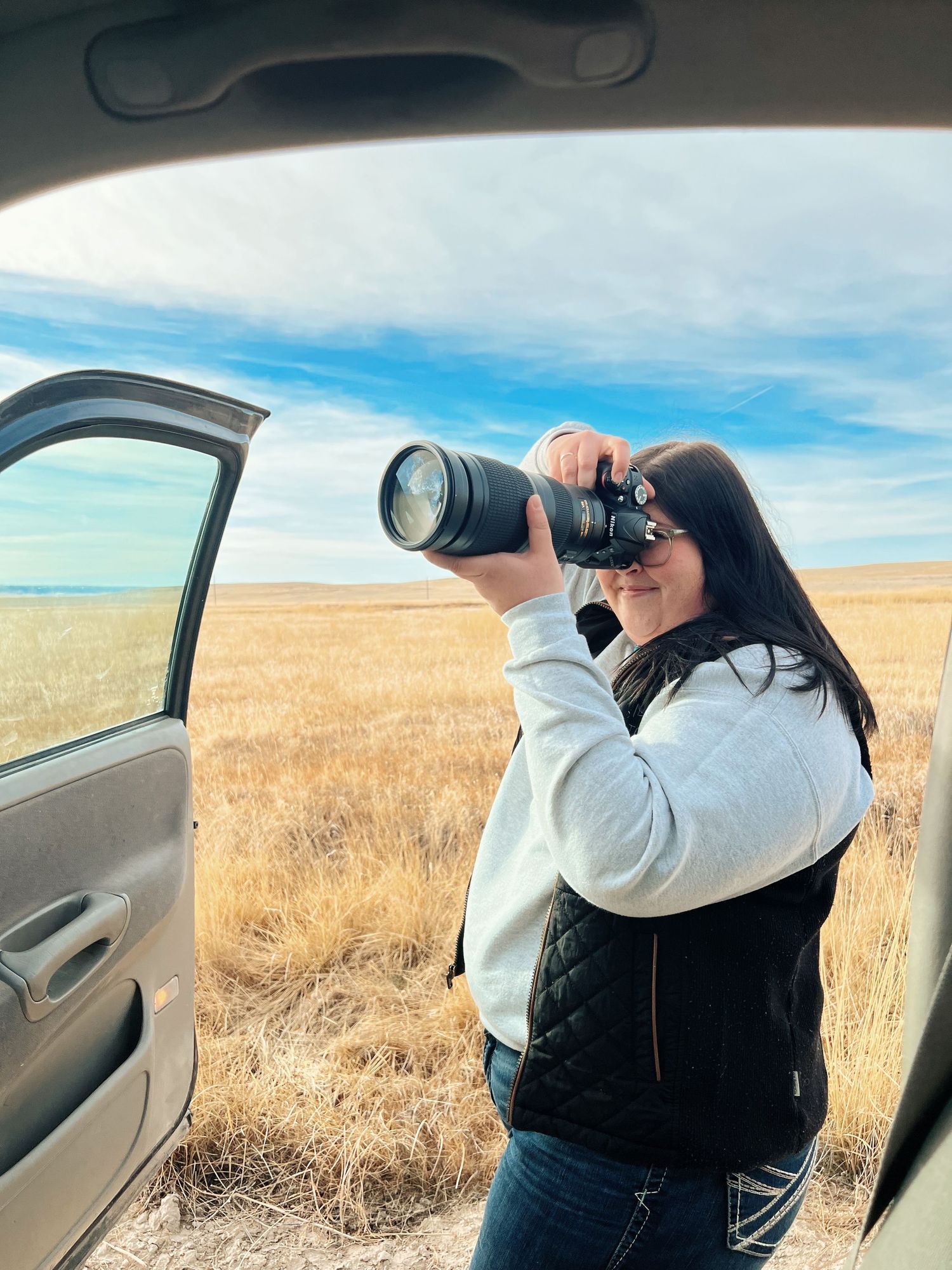 A photographer with a telephoto lens takes photos from a car door in a golden wheat field under blue skies.