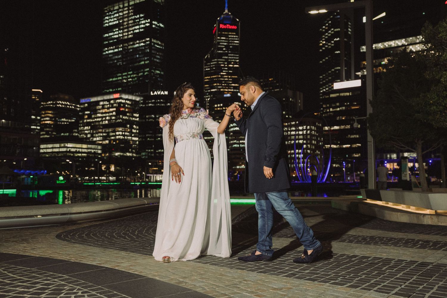 A couple walks along a city waterfront at night with illuminated skyscrapers in the background.